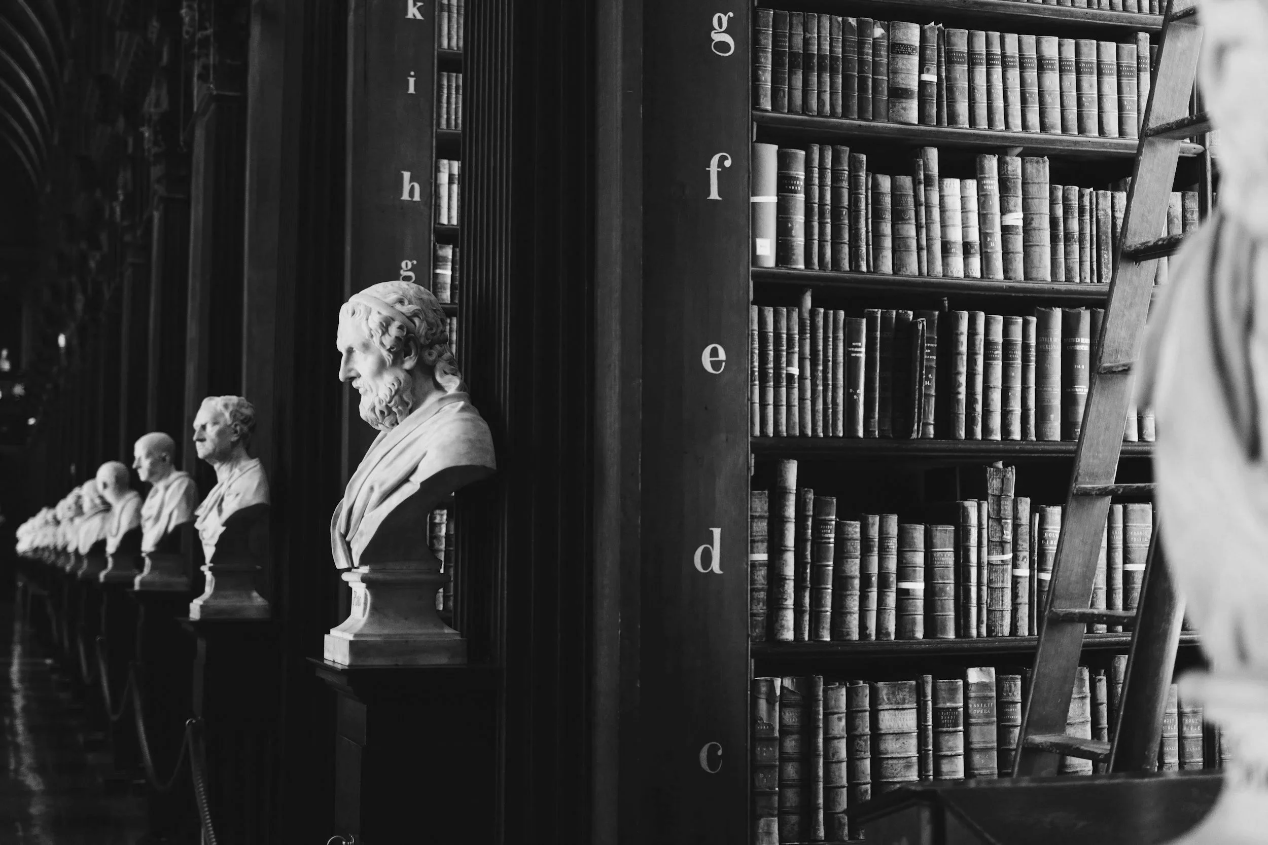Black and white photo of a library with shelves filled with old books. In front, there are bust statues of famous authors or historical figures, lined up on a table or shelf.