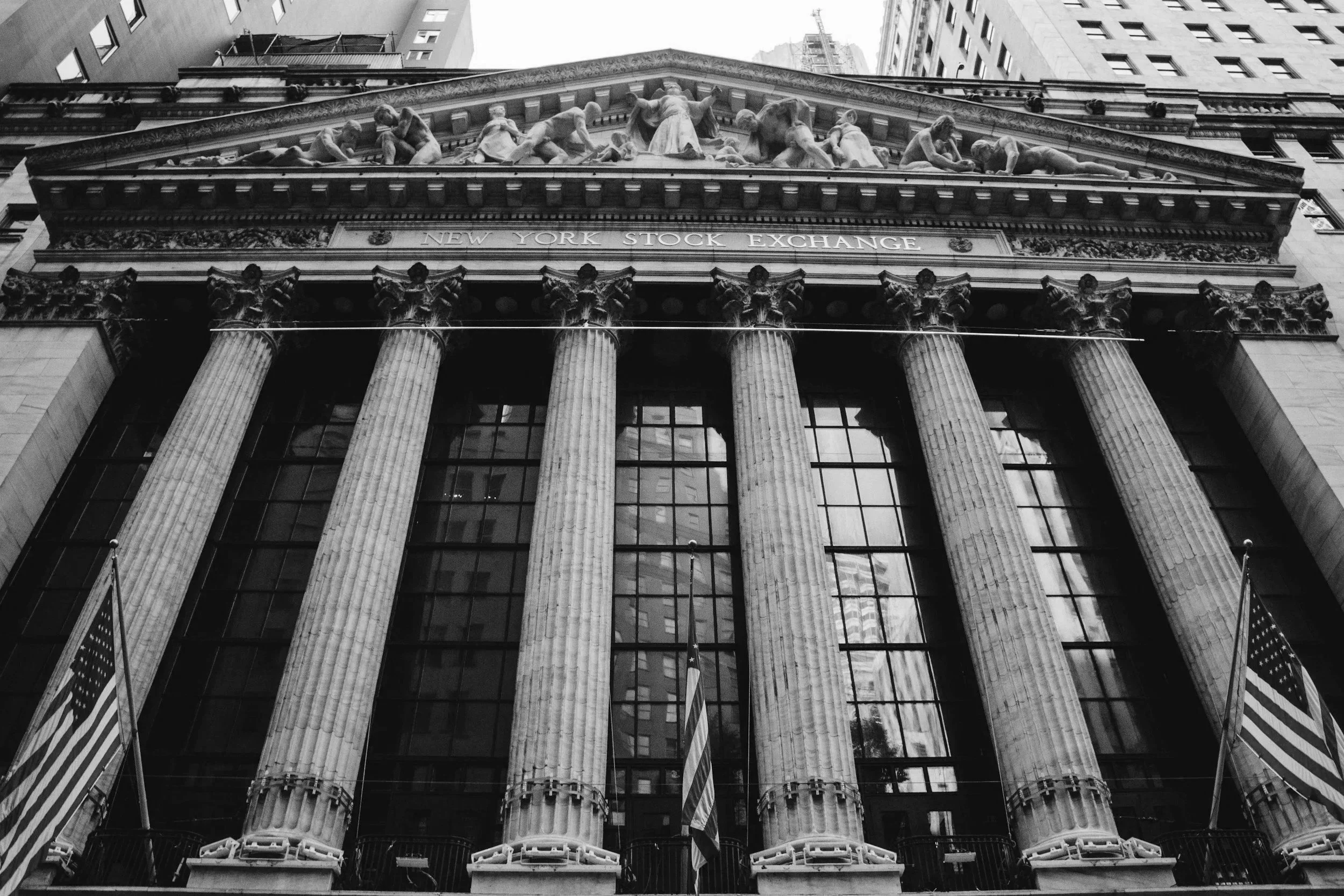 Black and white photo of the façade of the New York Stock Exchange building, with neoclassical columns and detailed sculptures on the pediment, surrounded by tall buildings.