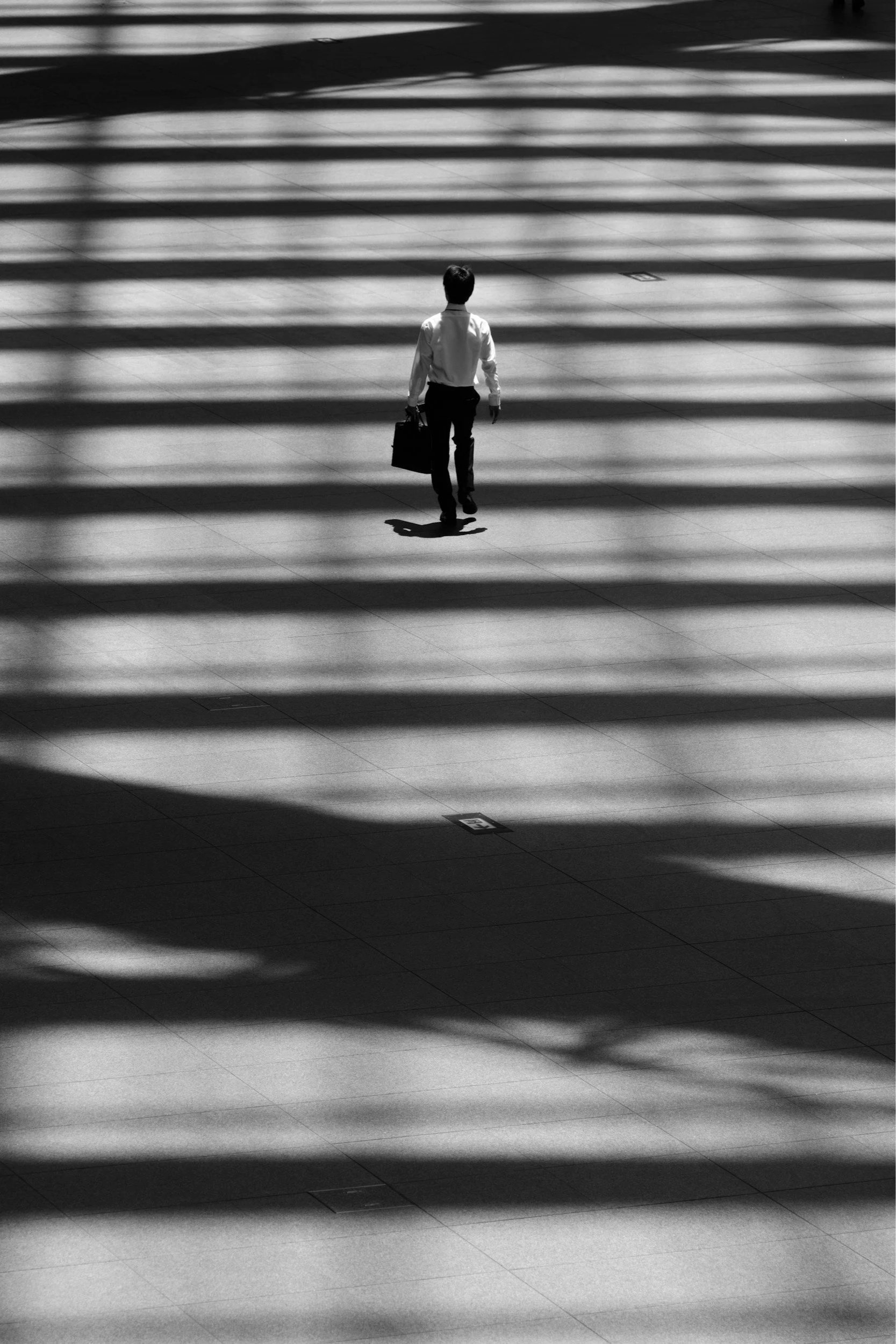 A man in business attire holding a briefcase walking through a modern building with sunlight casting linear shadows across the floor.