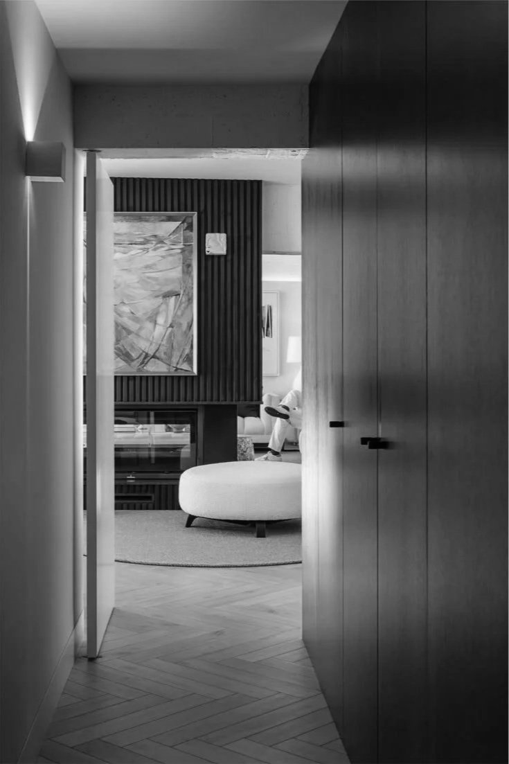 A narrow hallway leading into a modern living room with a round ottoman, a piece of abstract artwork, and a person sitting in the background.