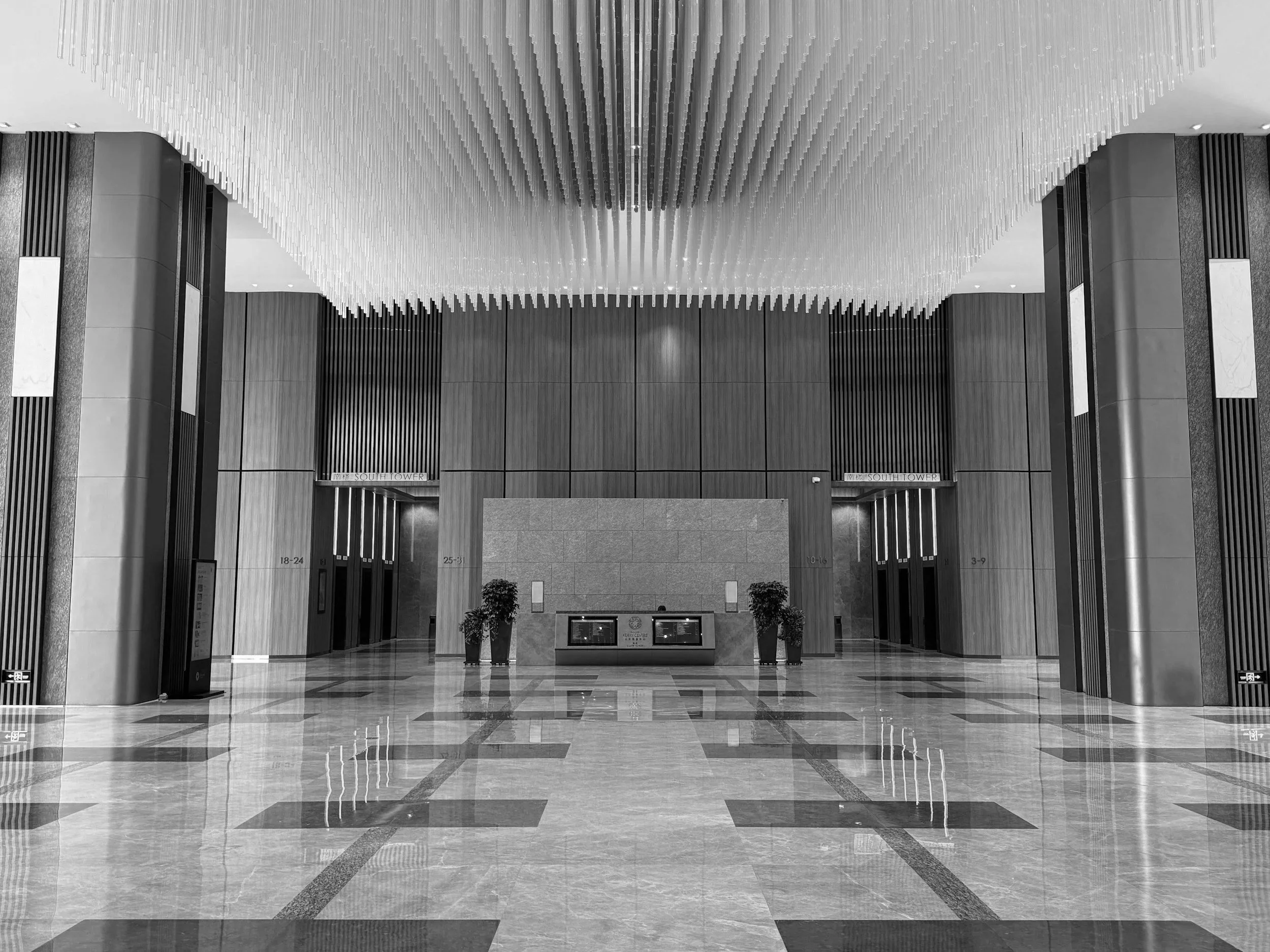 Interior view of a modern building lobby with polished stone floors, wooden wall panels, and decorative pillars. There is a reception desk in the center with two potted plants and a large wall behind it. Multiple elevators are visible on both sides of the room.