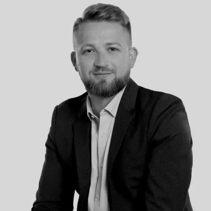 Black and white professional portrait of a young man with a beard, wearing a suit and collared shirt, smiling slightly, against a plain background.