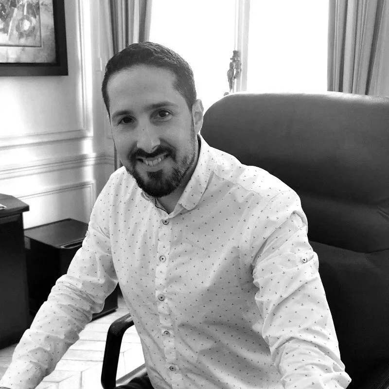 A man with a beard and short hair, smiling, seated in a black office chair in a room with curtains and framed artwork.