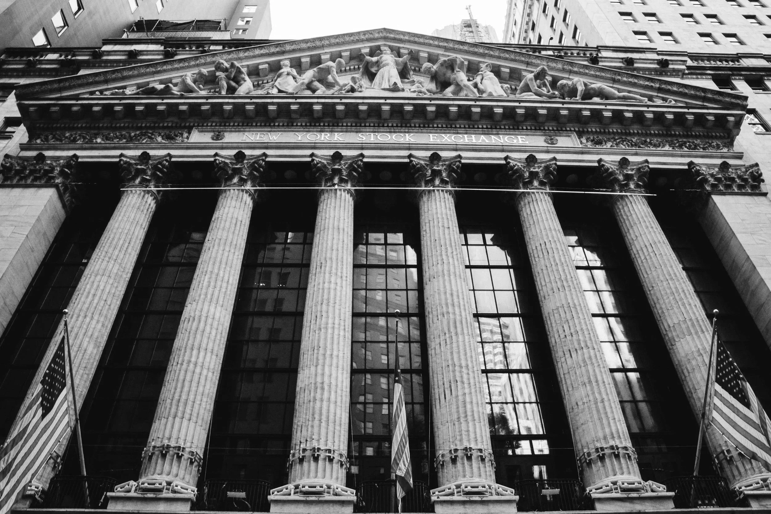 Front view of the New York Stock Exchange building with tall columns and classical sculptures at the top, city buildings in the background, black and white photograph.