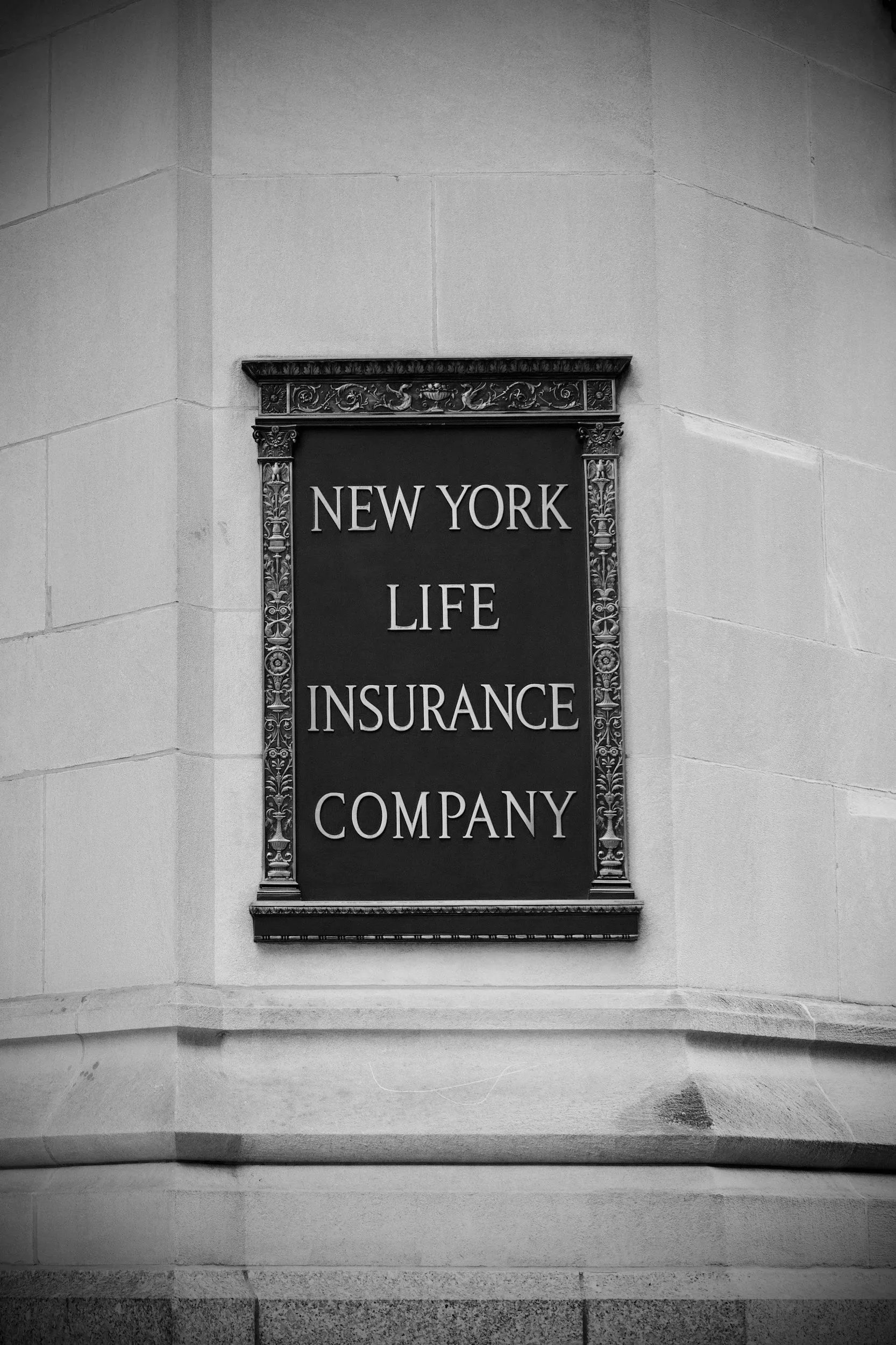 A black and white signboard on a stone wall reading 'New York Life Insurance Company' with ornate decorative framing.