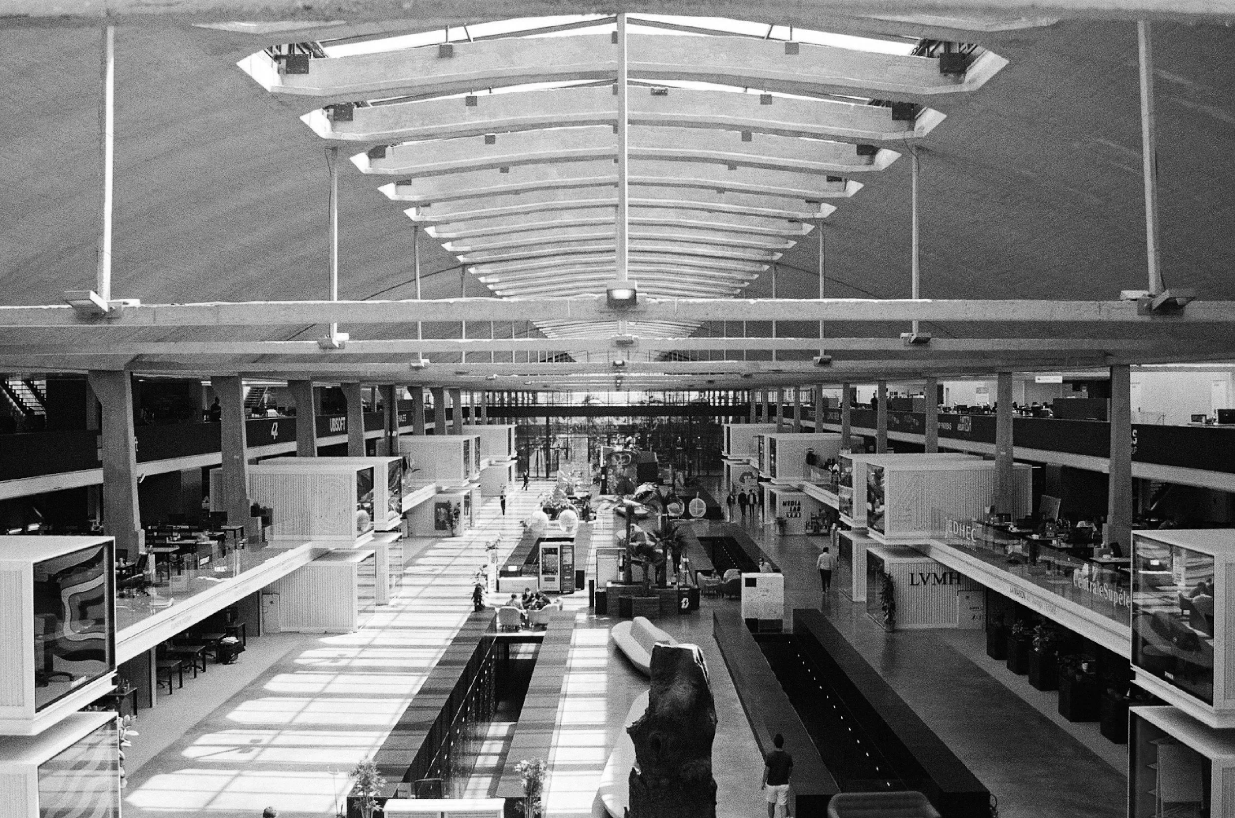 Black and white photo of an indoor shopping mall or airport terminal with multiple levels, escalators, and shops. People are walking throughout the space.