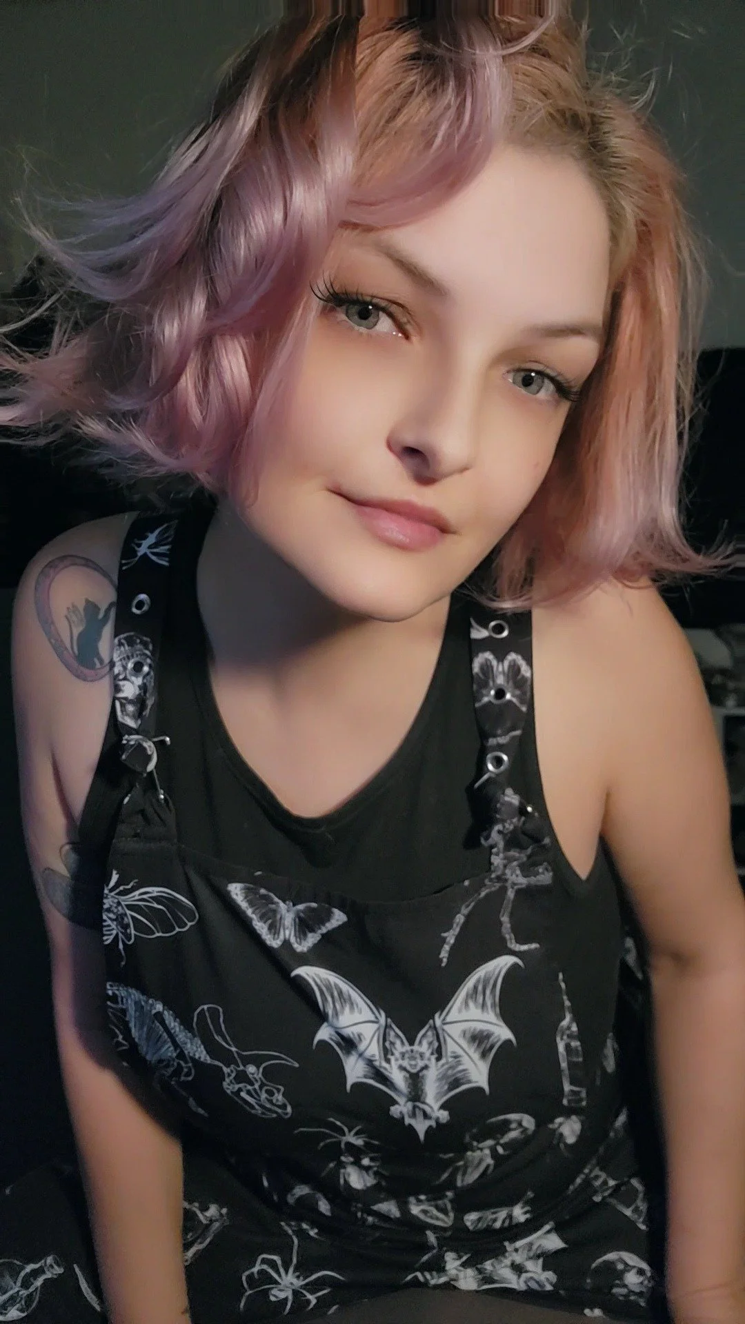 Young woman with pink and purple wavy hair, wearing a black sleeveless top with bat, butterfly, skull, and other gothic-themed designs, sitting indoors.