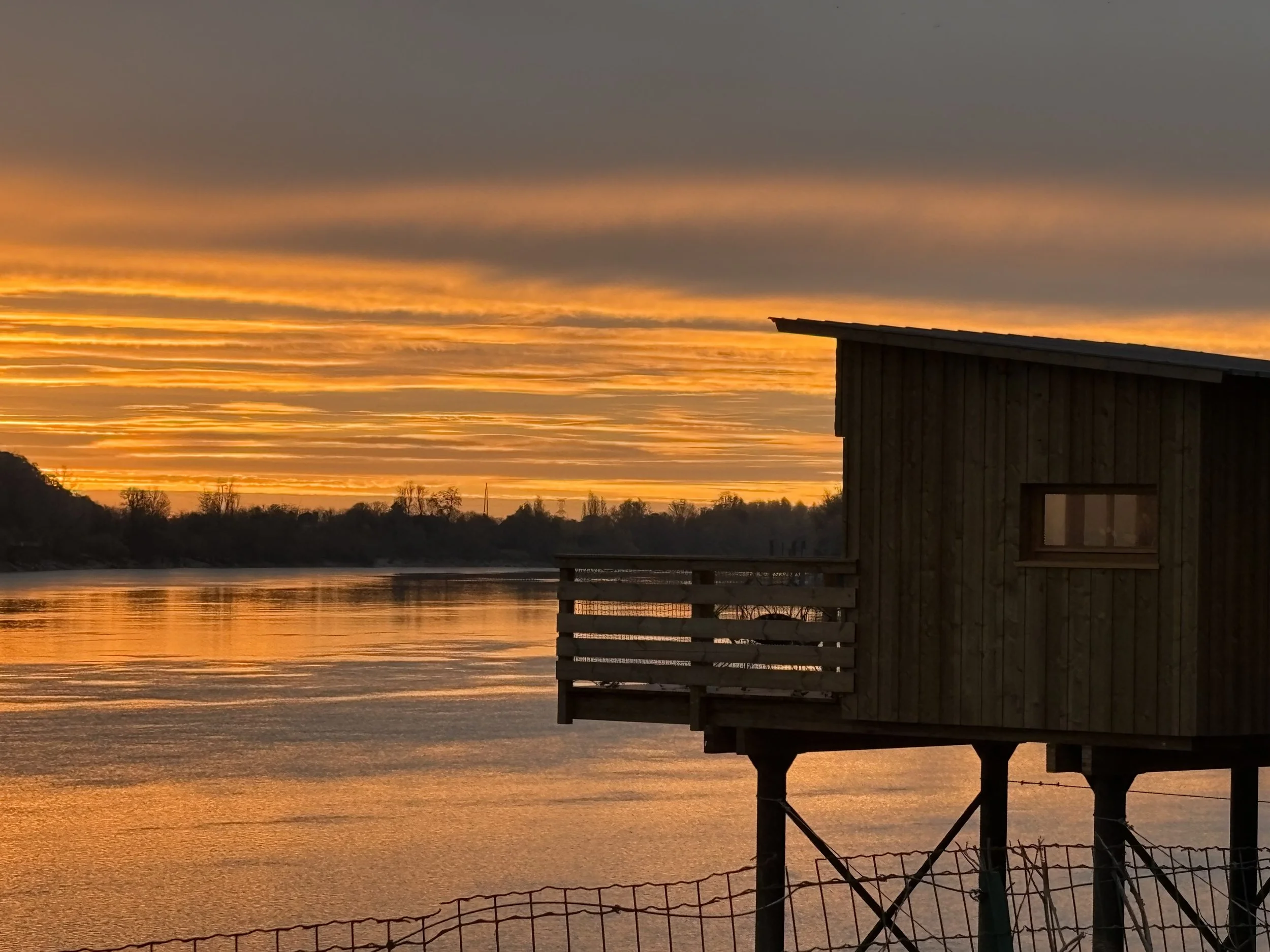 Cabane en bois sur pilotis au bord d'un lac ou d'une rivière au coucher du soleil, avec un ciel orageux et des nuages colorés.