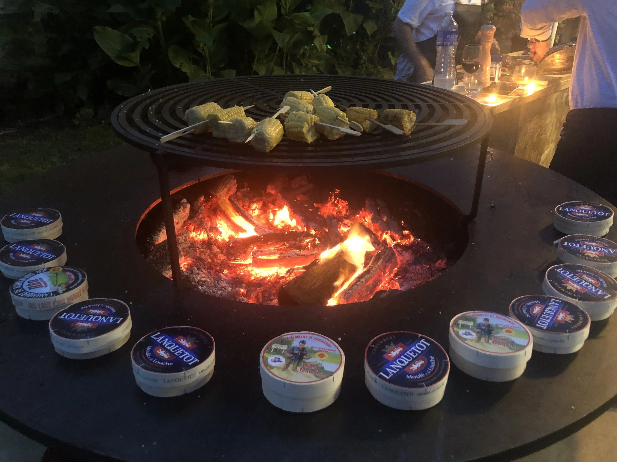 Cornichons en brochettes sur un grill au-dessus d'un feu de bois, entouré de pots de moutarde Lancouet, lors d'une soirée en plein air.
