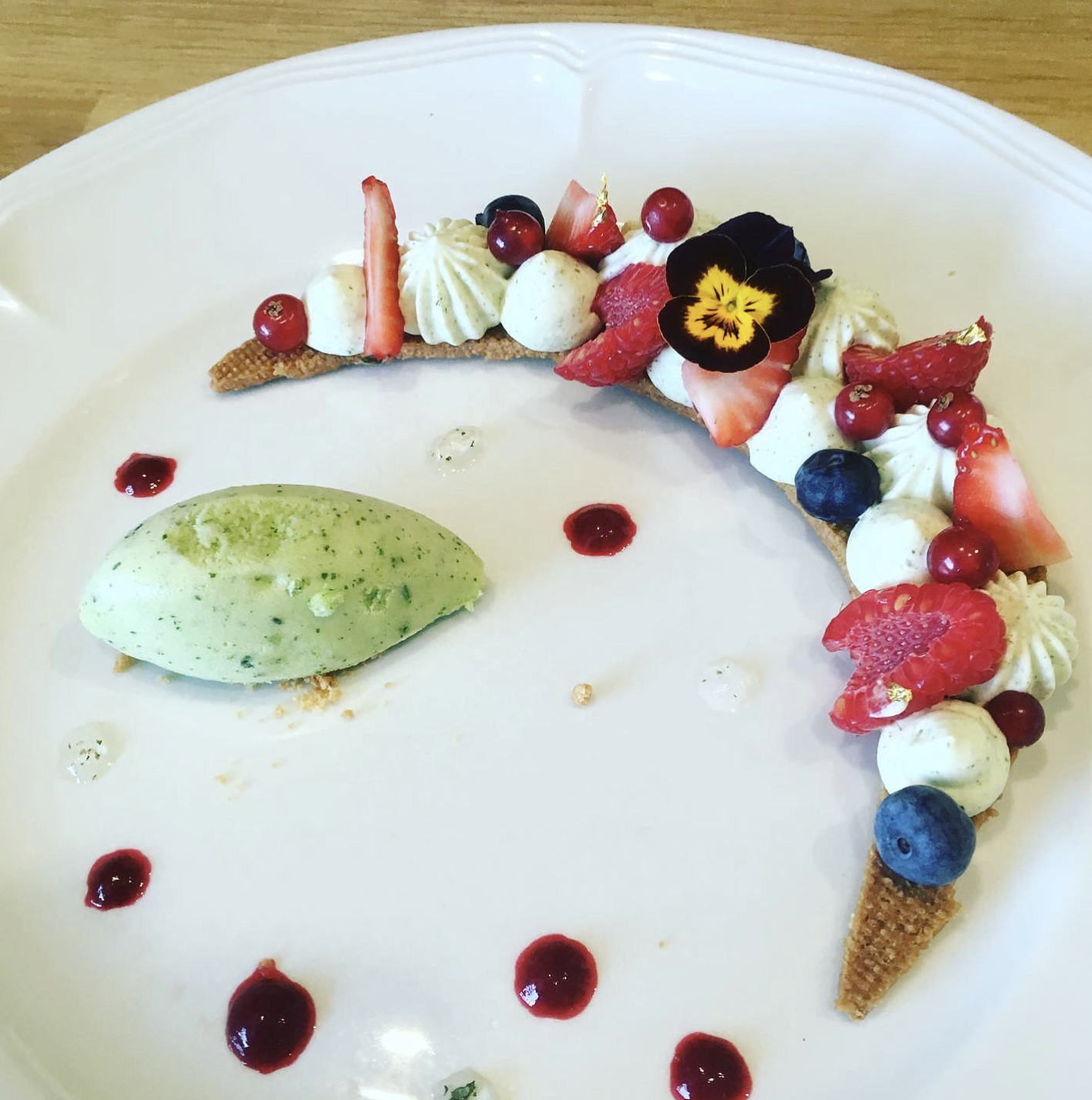Gâteau en forme de croissant orné de fruits rouges, myrtilles, pistaches, et fleurs, avec une boule de glace à la pistache à côté, sur une assiette blanche.