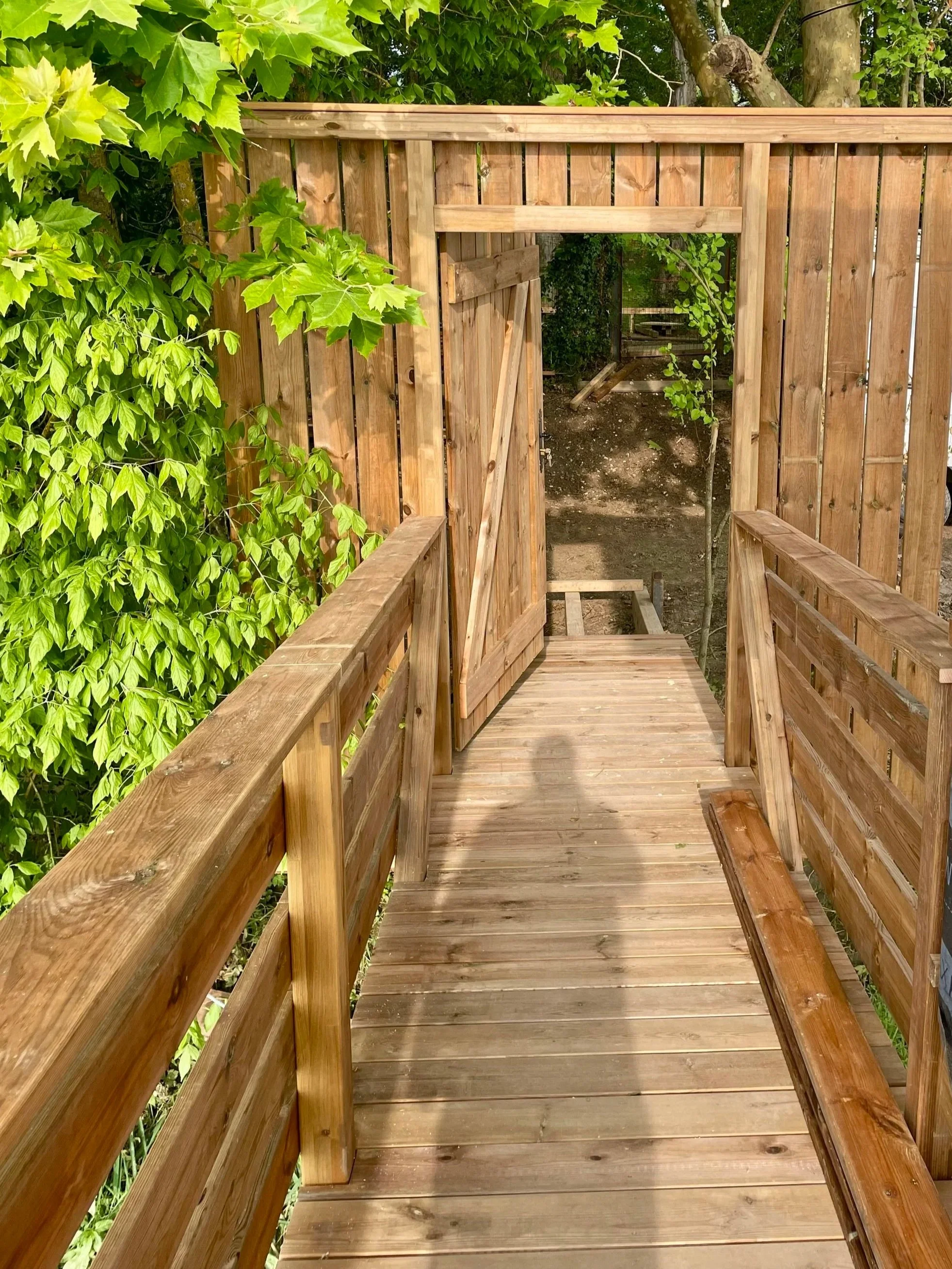 Passerelle en bois avec clôtures en bois, entrouverture menant à une zone extérieure plantée d'arbres, de feuillage et de terre.