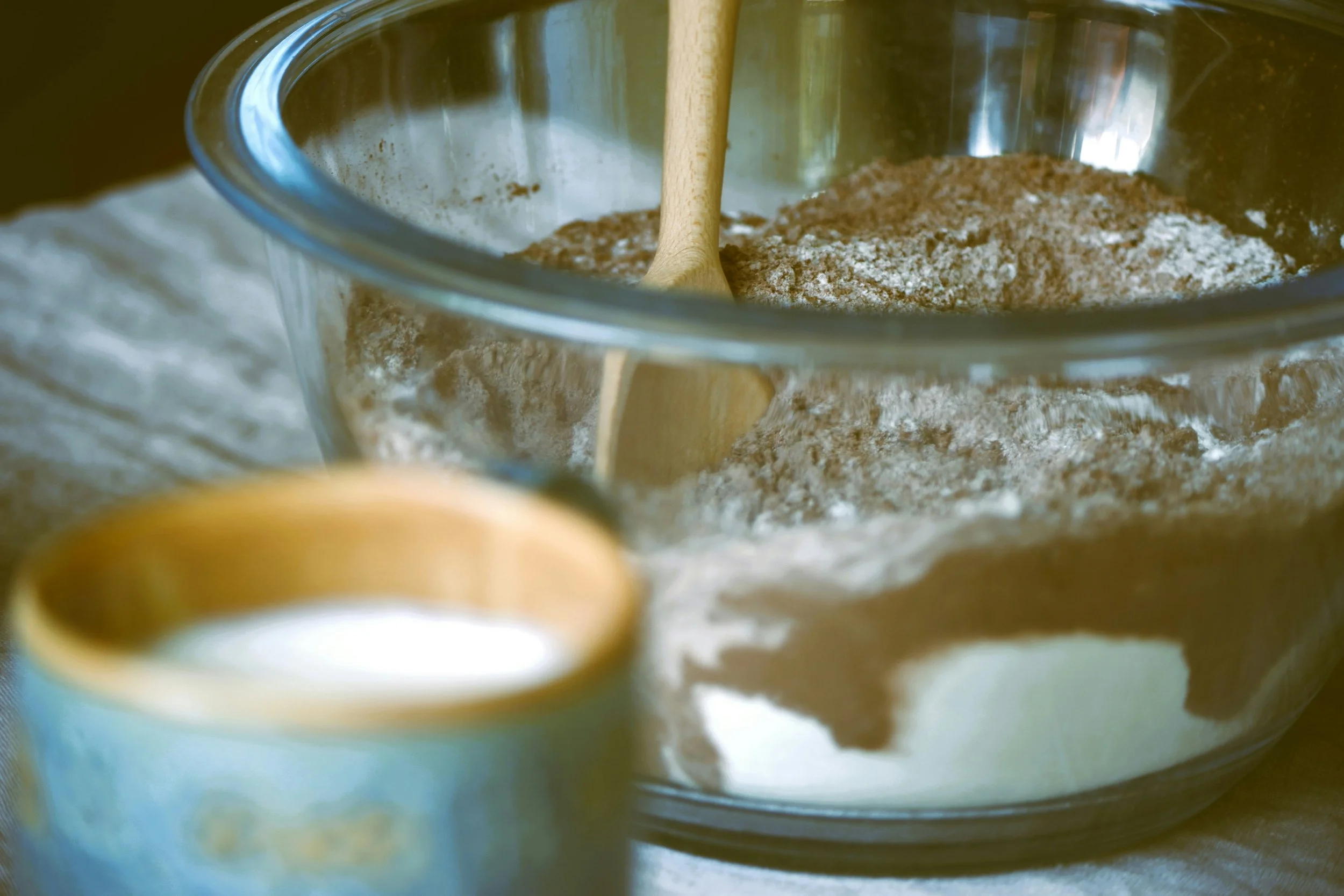 Un bol en verre contenant de la farine, avec une cuillère en bois à l'intérieur, placé sur une surface en tissu. Une tasse en céramique dorée et bleue en premier plan.