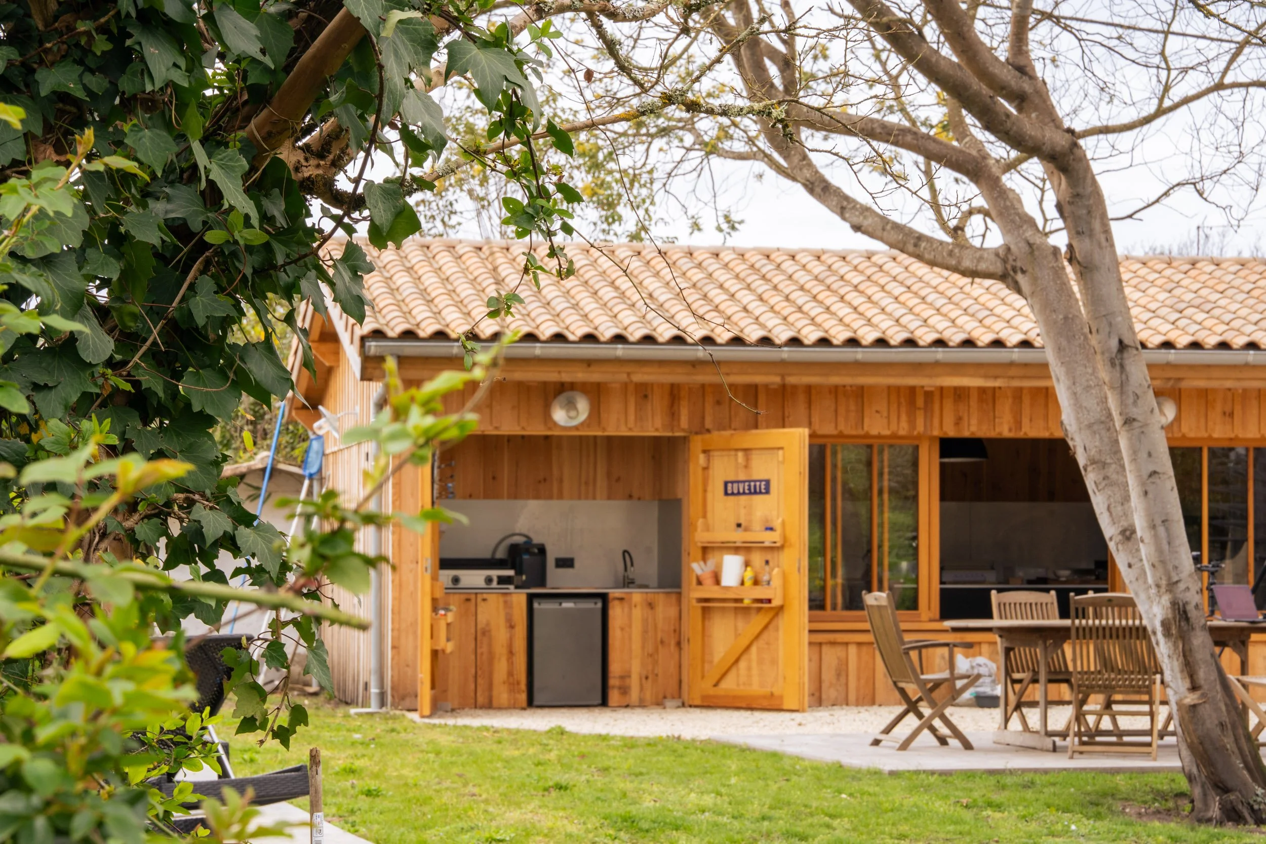 Une cabane en bois avec un toit en tuiles, une terrasse avec une table et des chaises, et un espace cuisine extérieur dans un jardin avec des arbres.