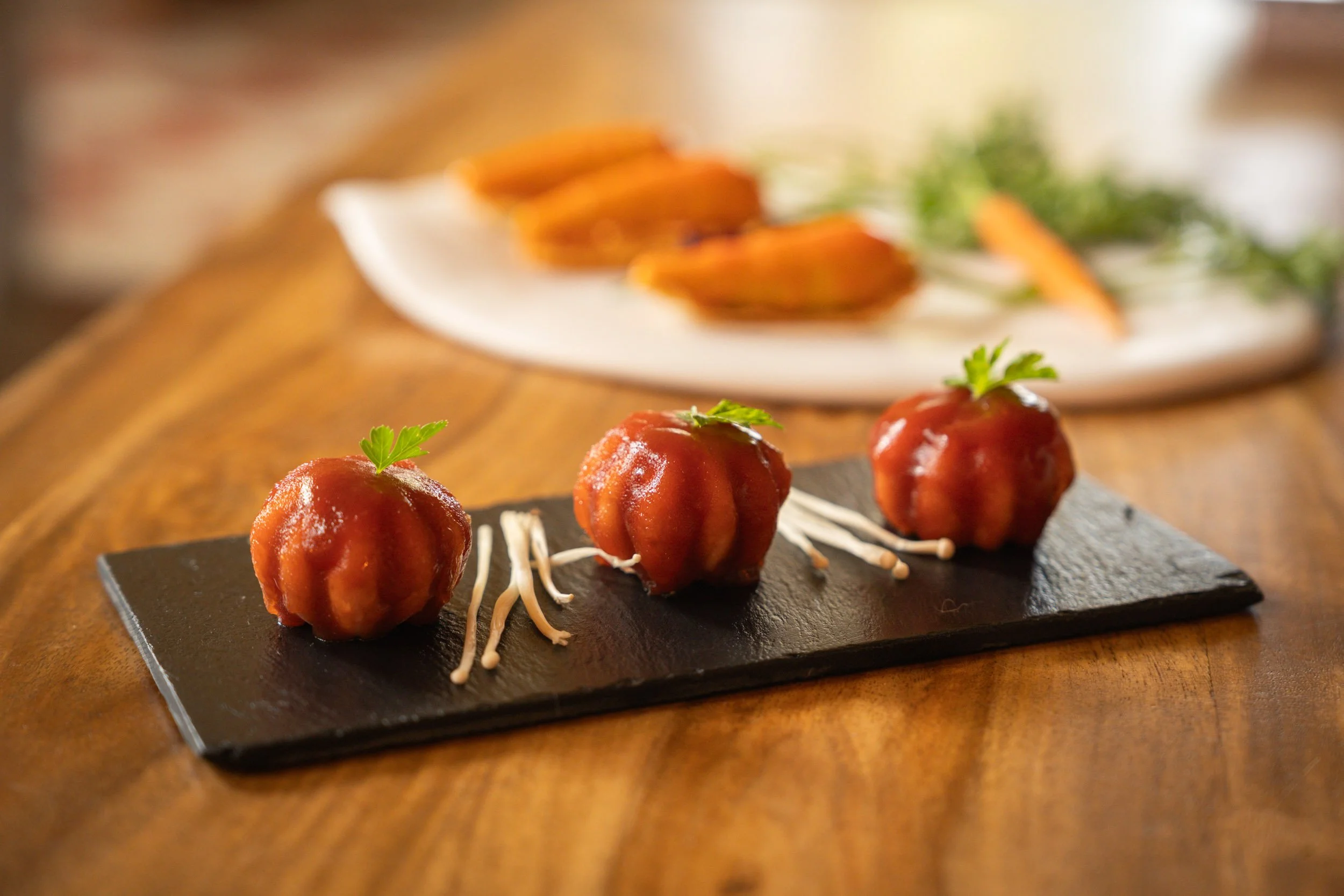 Trois petites boules de viande ou de légumes en sauce sur une planche en ardoise, garnies de jeunes pousses de coriandre, avec des pousses de soja en dessous. En arrière-plan, une assiette blanche avec des morceaux de patate douce cuite ou friture.