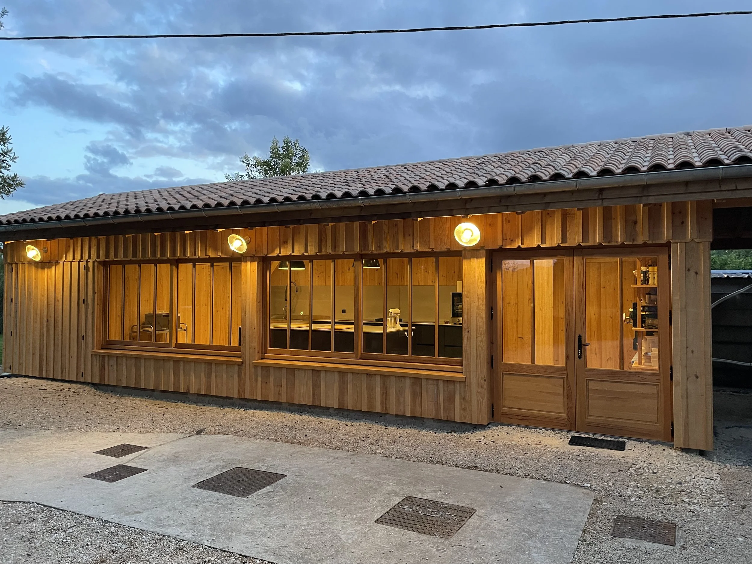 Une maison en bois éclairée la nuit, avec de grandes fenêtres, une porte en bois et un toit en tuiles, sous un ciel nuageux.