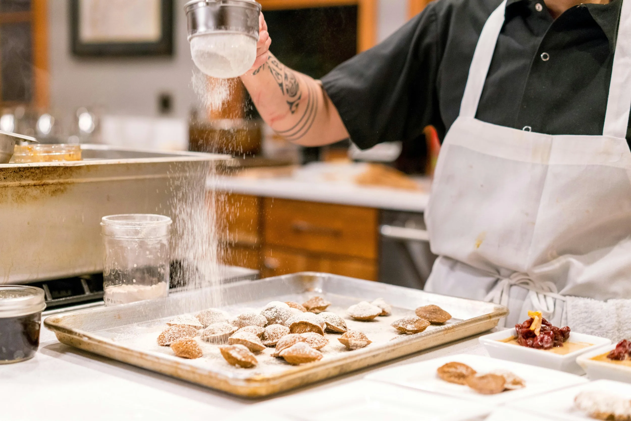 Une personne en tablier blanc prépare des biscuits en saupoudrant de la farine, avec plusieurs biscuits cuits sur une plaque, dans une cuisine.