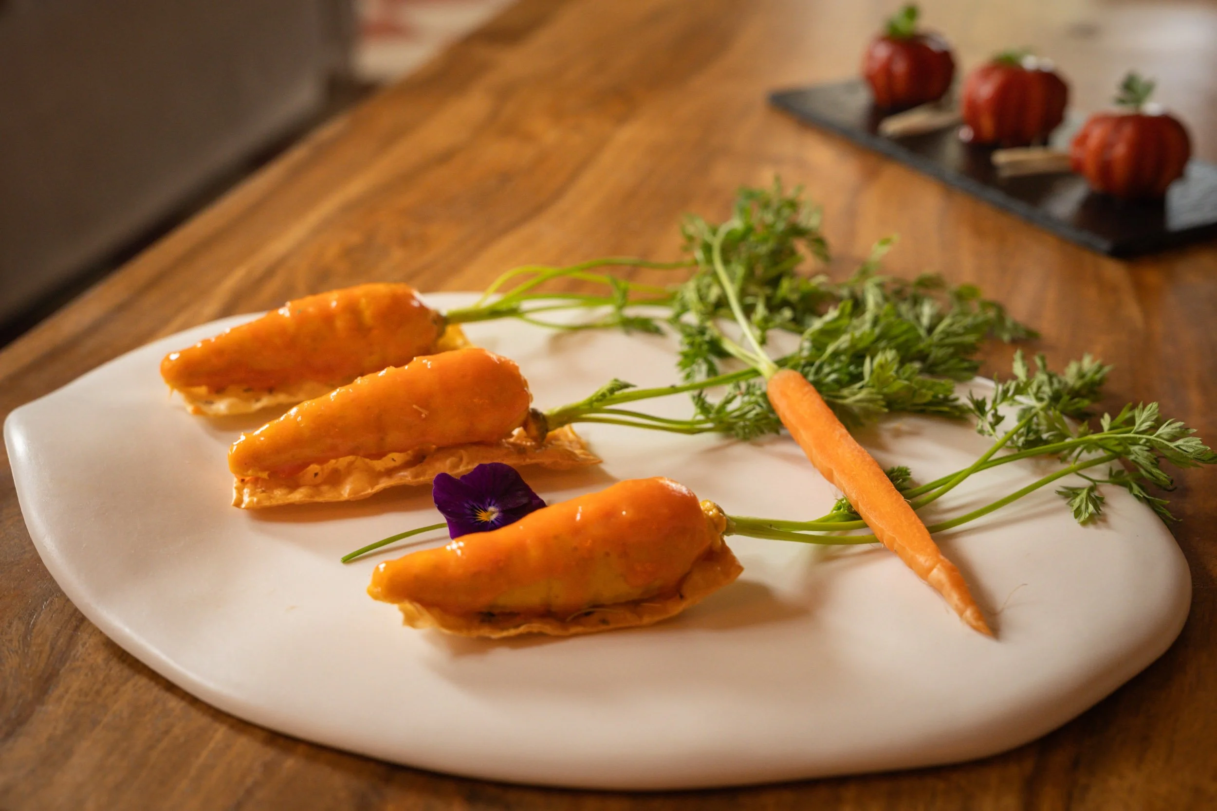 Assiette avec plusieurs aubergines farcies, une carotte, des carottes en fleurs comestibles, et des herbes, sur une table en bois.