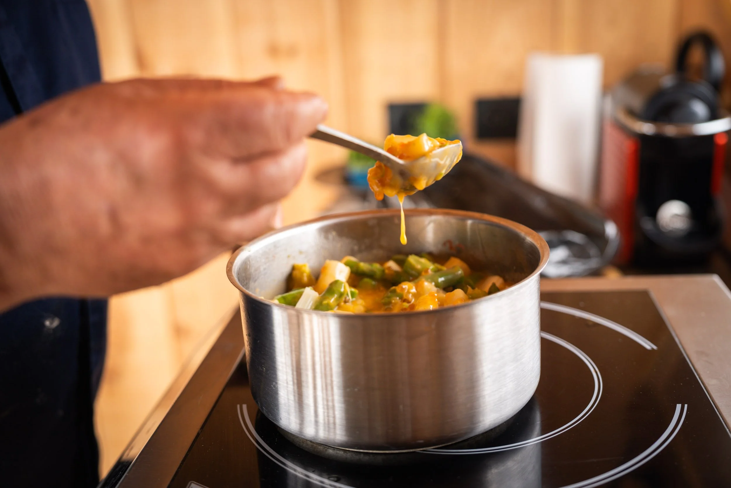 Une personne touche une casserole contenant une soupe avec des légumes verts et jaunes sur une plaque de cuisson électrique.