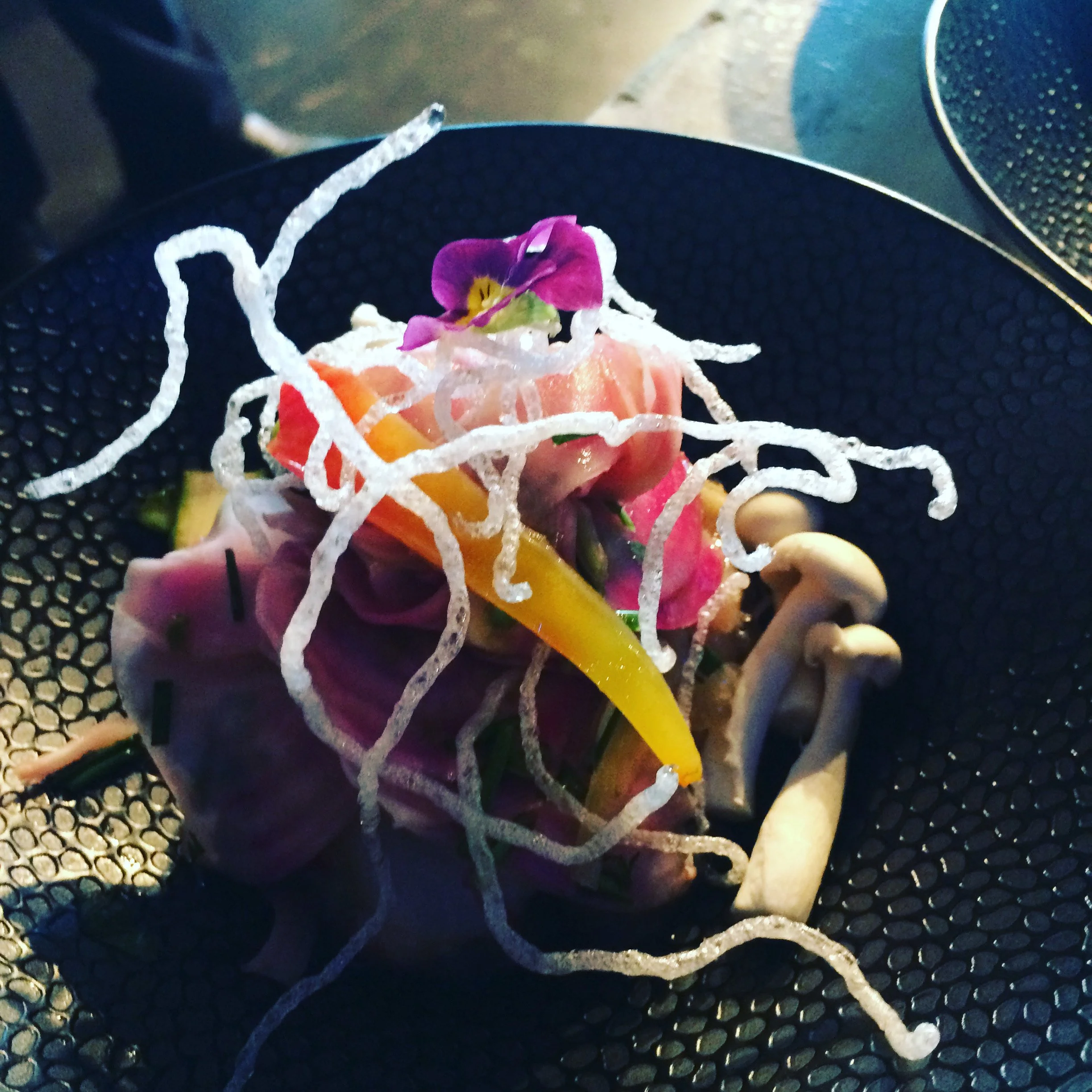 Salade composée avec des légumes colorés et des champignons, décorée de fleurs violettes et de filaments croustillants.