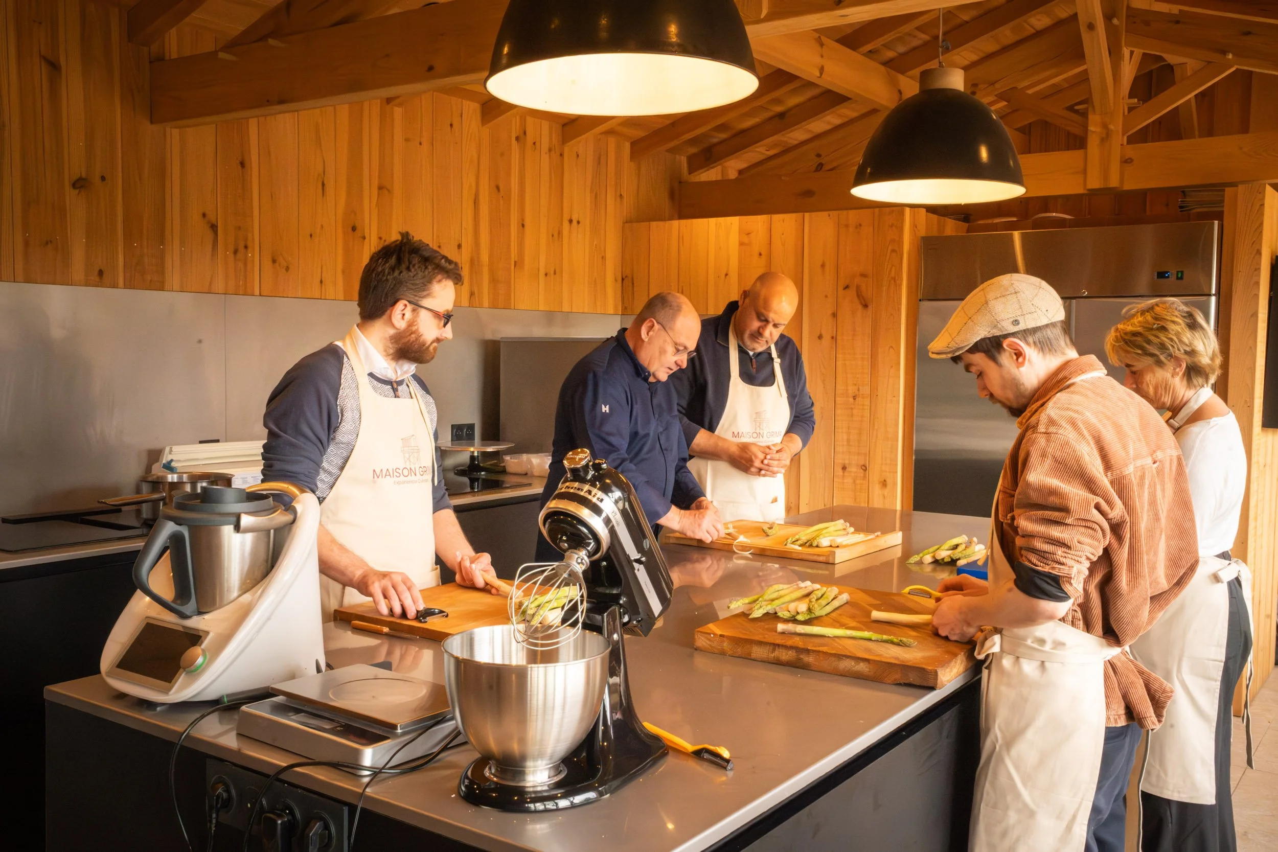 Groupe de cinq personnes en train de cuisiner dans une cuisine en bois, portant des tabliers, préparant des asperges sur des planches à découper.