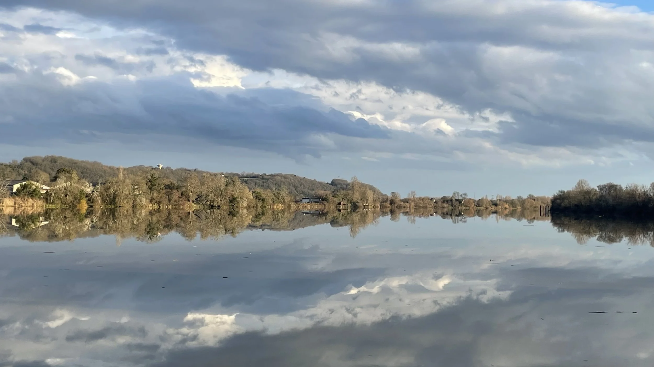 Un lac calme reflétant les nuages et le ciel au-dessus, avec des arbres et quelques bâtiments en arrière-plan.