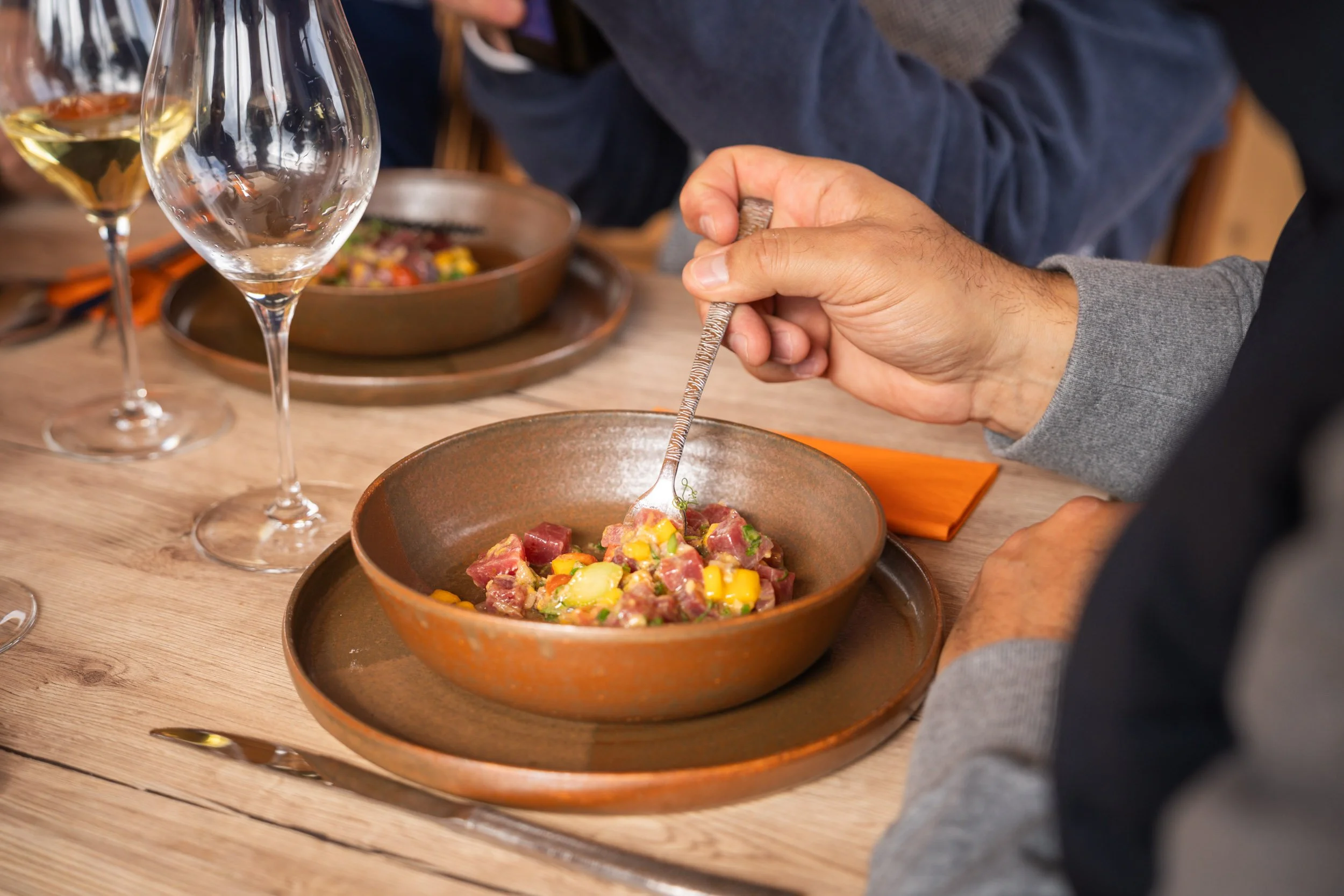 Gastronomie dans un restaurant : personne mangeant un tartare de viande avec des légumes, autres verres à vin sur la table en bois.