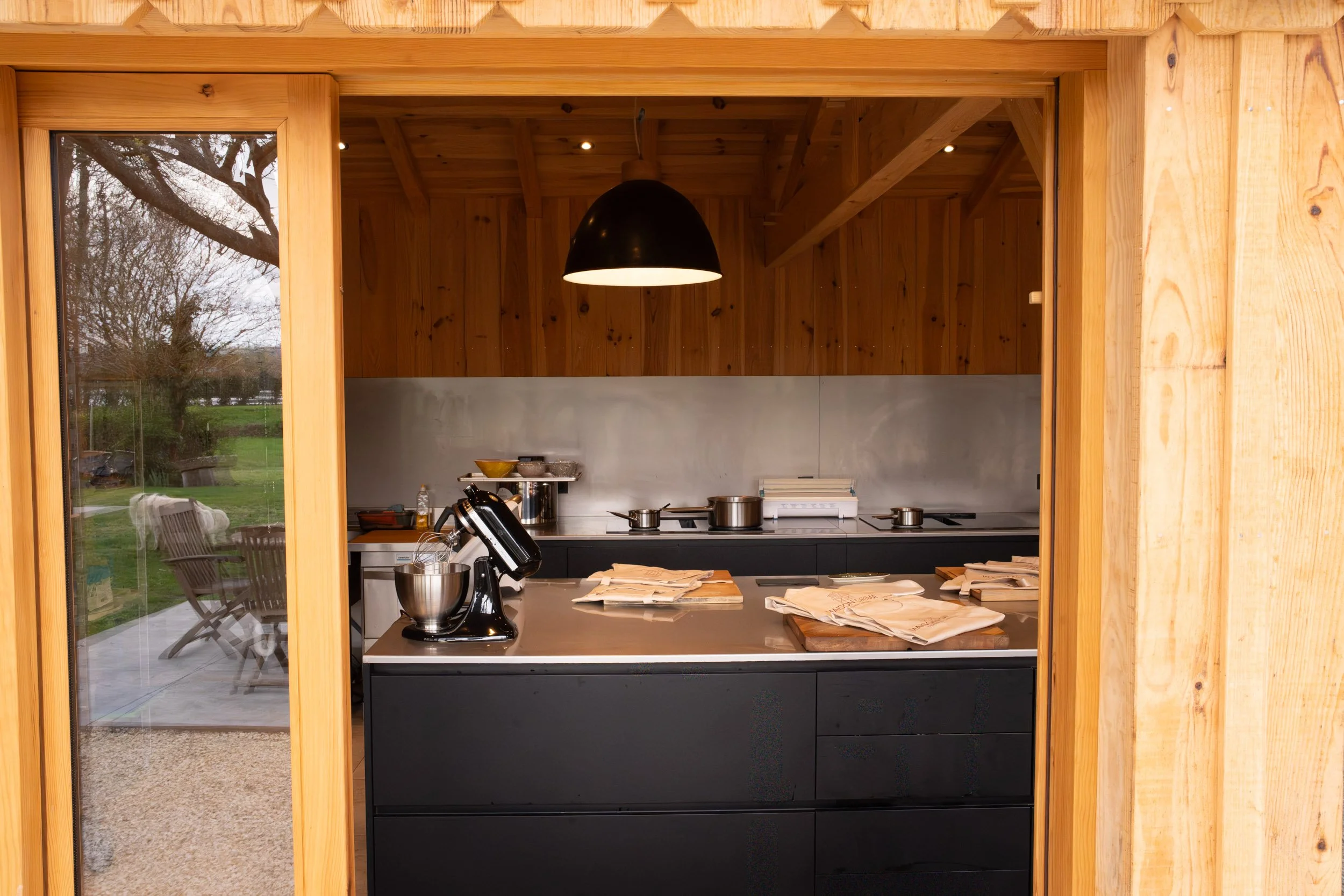 Cuisine moderne avec plan de travail noir, étagères en bois, lampadaire noir suspendu, et vue à l'extérieur à travers une porte vitrée.