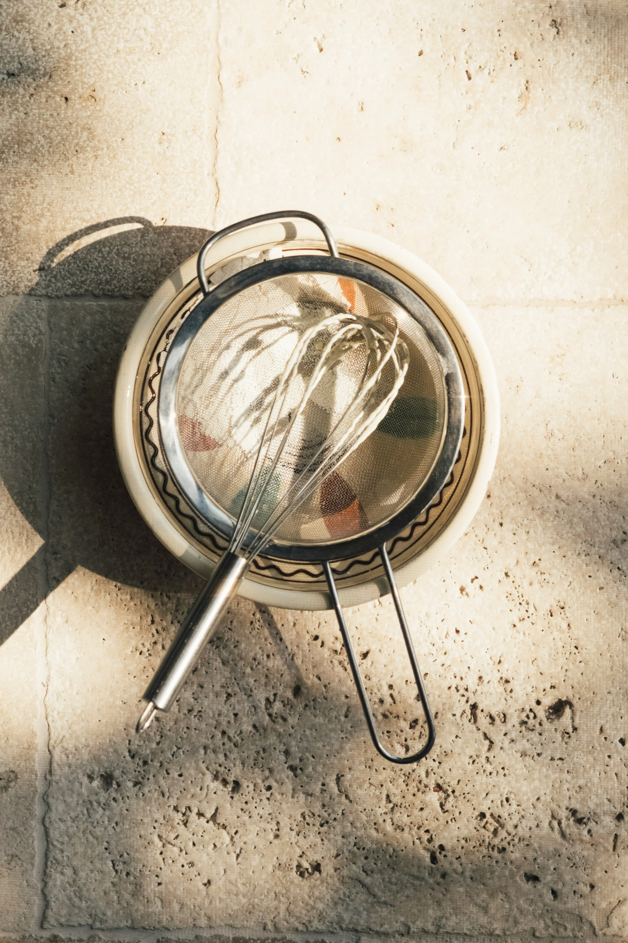Une passoire avec un fouet à mélanger dessus, vue d'en haut, sur un sol en pierre.
