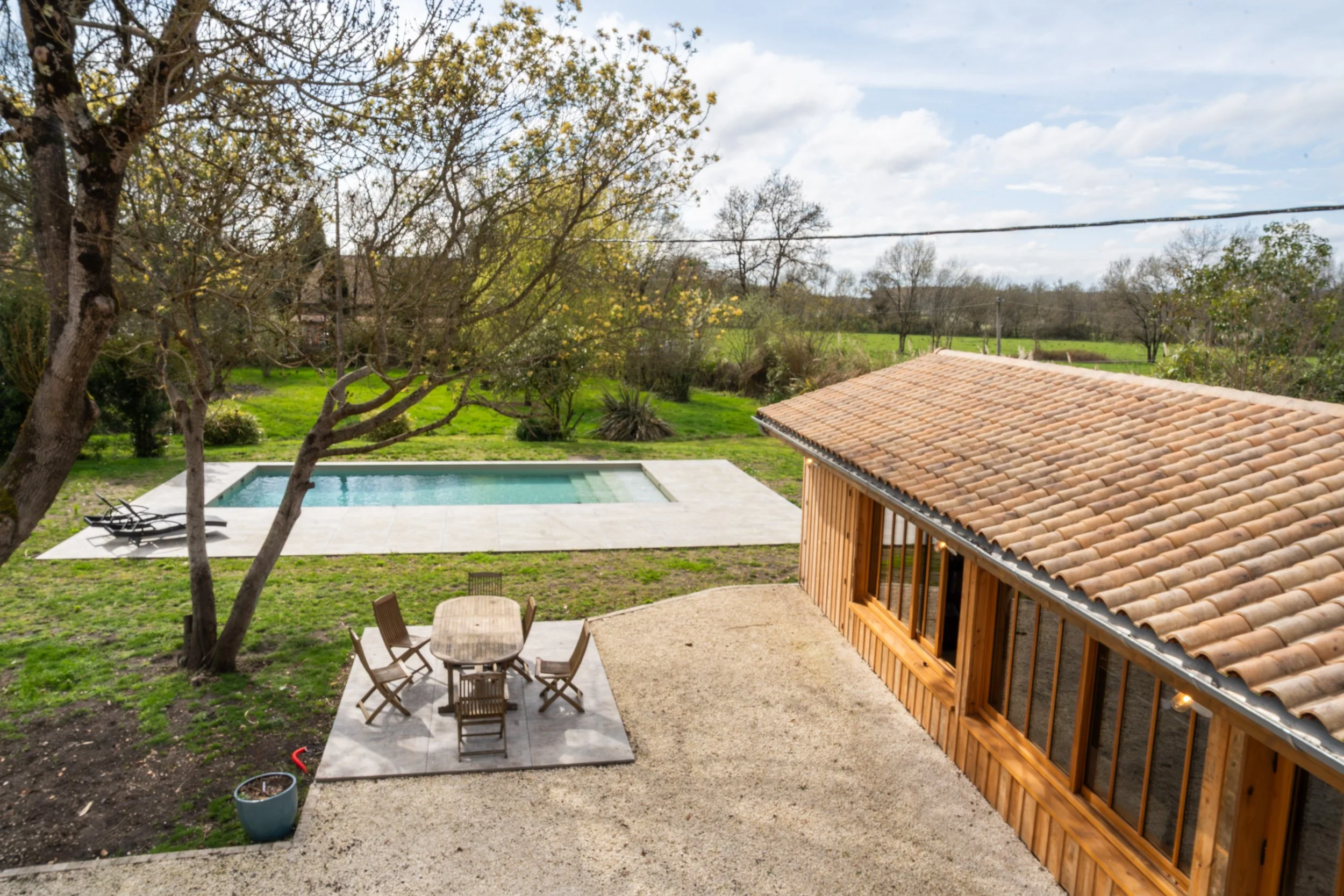 Une terrasse avec un espace repas en bois, une piscine dans un jardin verdoyant entouré d'arbres, et une maison en bois avec un toit en tuiles.