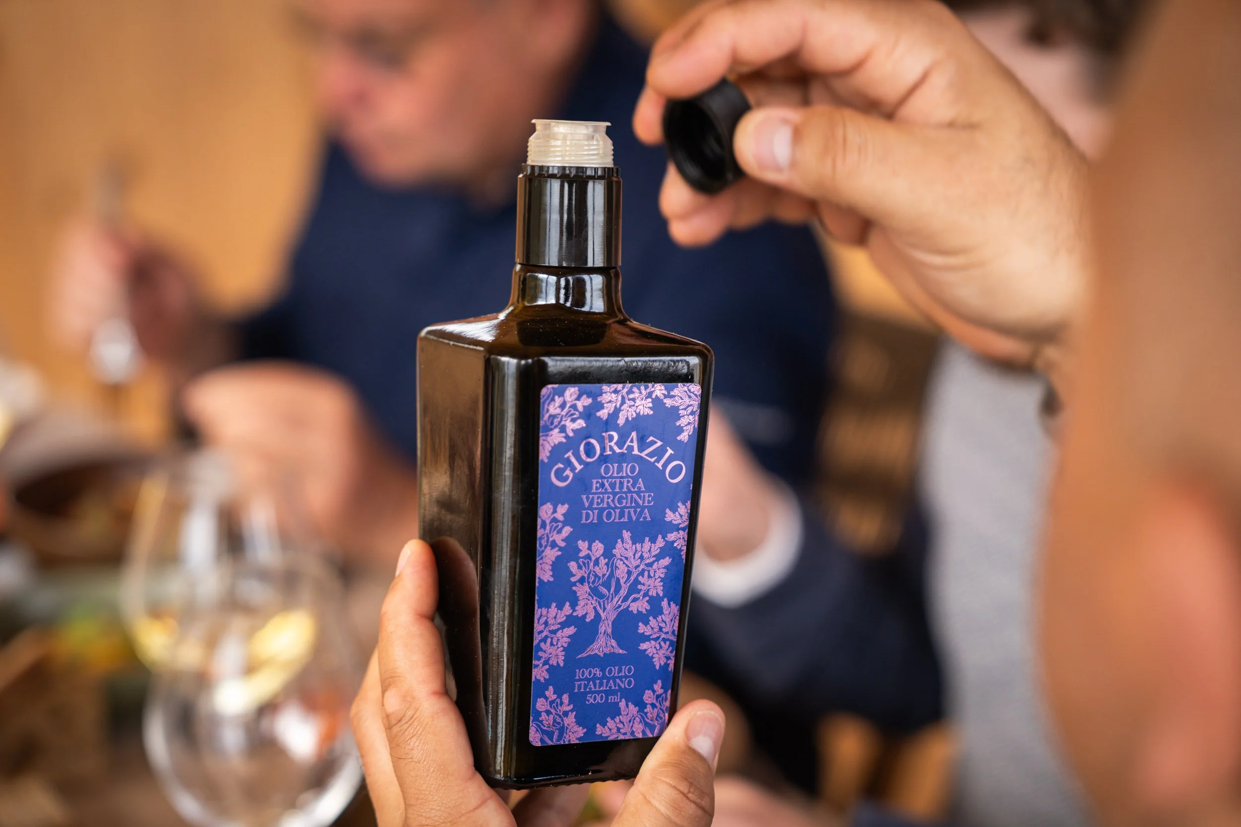 Une personne tient une bouteille d'huile d'olive italienne, marque GiORAZIO, avec un distributeur noir, dans un cadre de repas avec d'autres personnes floues en arrière-plan.