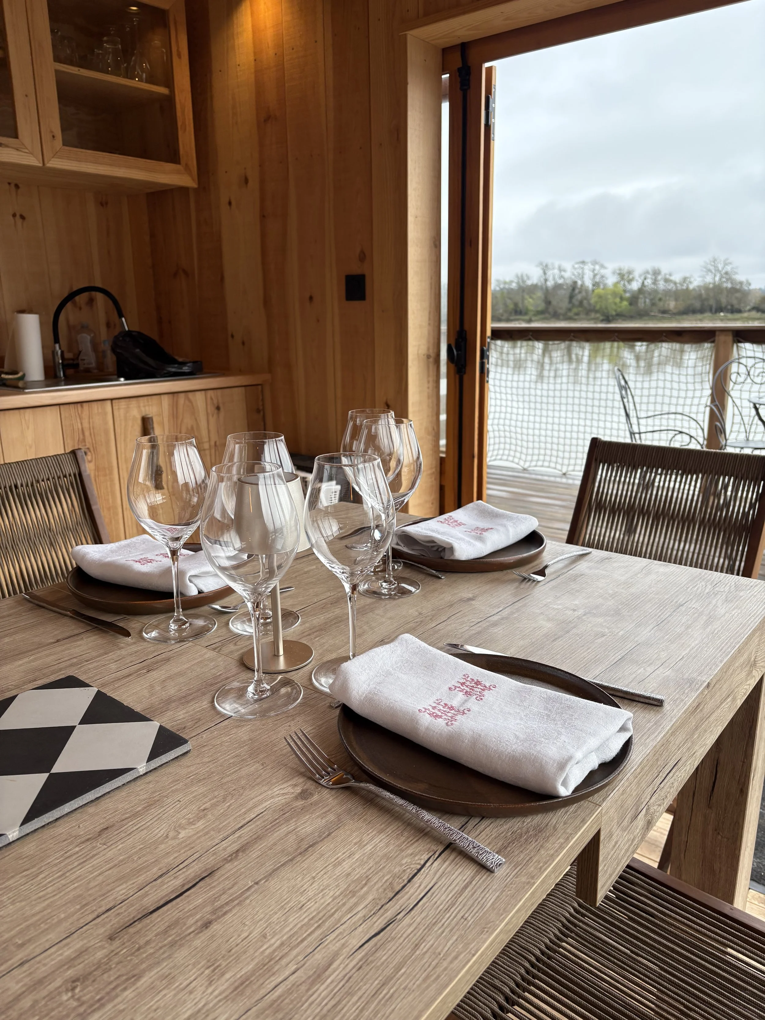 Une table de restaurant en bois est dressée avec des assiettes, des verres à vin, des couverts, et des serviettes blanches avec des motifs rouges. Une porte en bois s'ouvre sur une terrasse avec vue sur un lac et des arbres en arrière-plan.