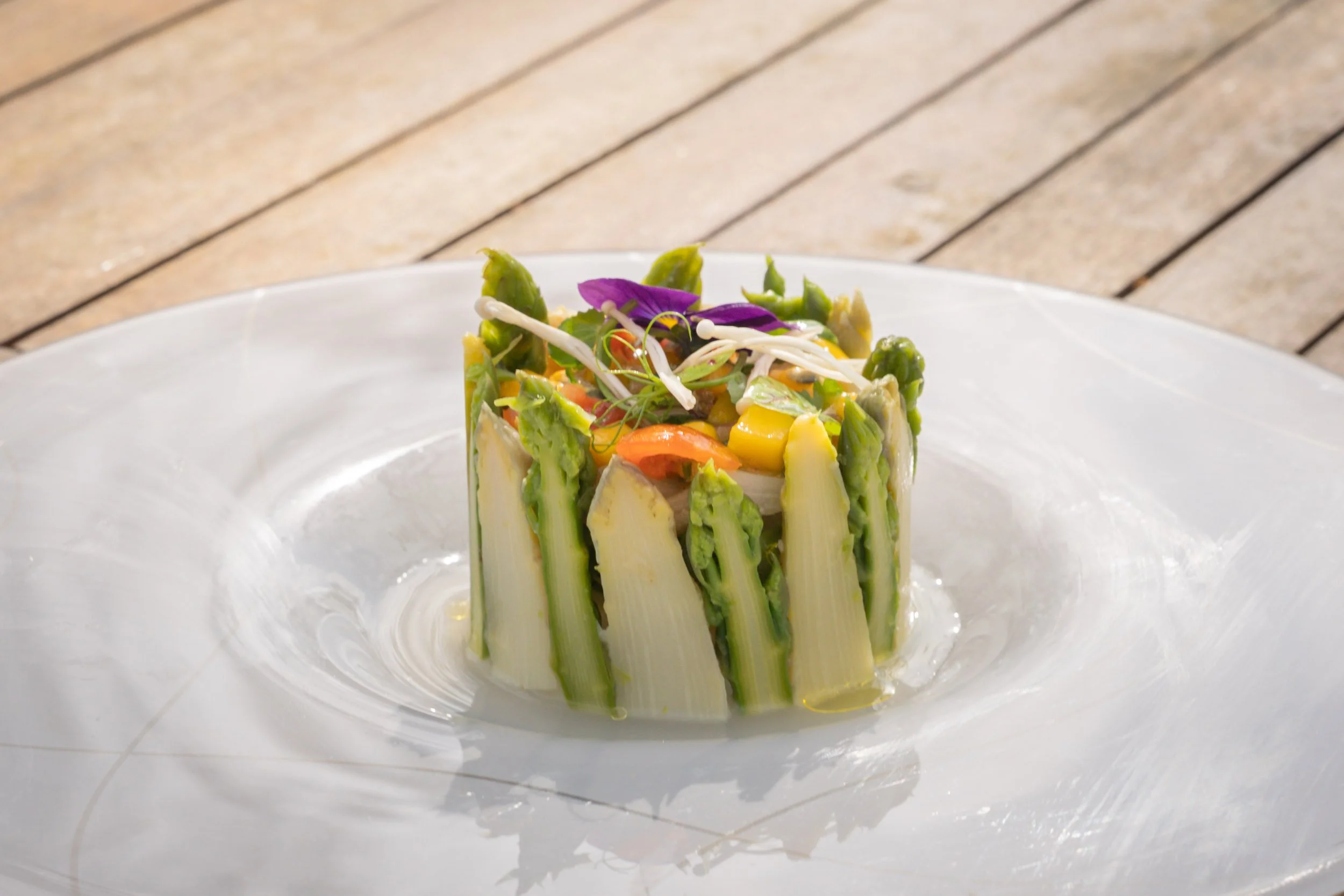 Une création culinaire avec des asperges et des légumes, décorée avec des micro-pousses et des fleurs comestibles, présentée dans un cercle sur une assiette blanche.