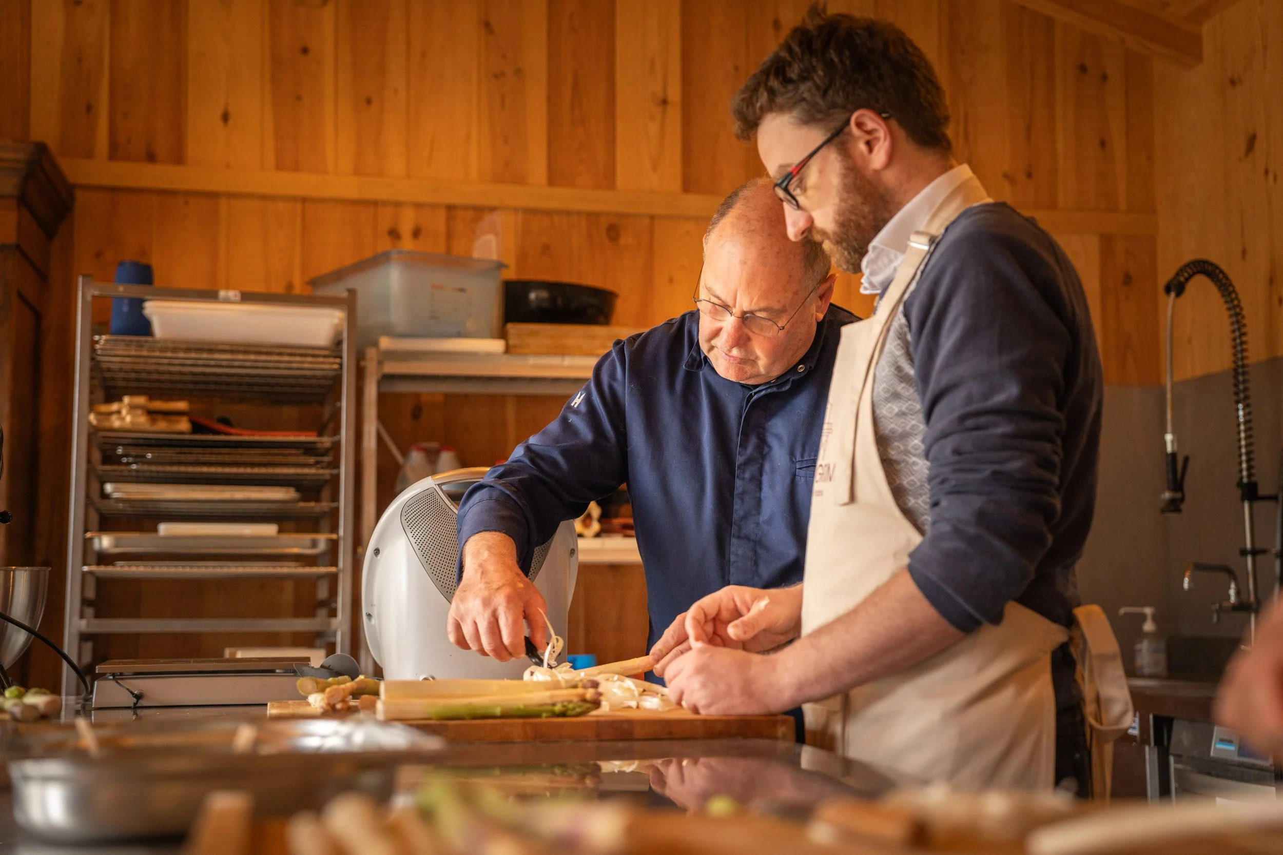 Deux hommes cuisinant ensemble dans une cuisine en bois, concentrés sur la préparation des aliments.
