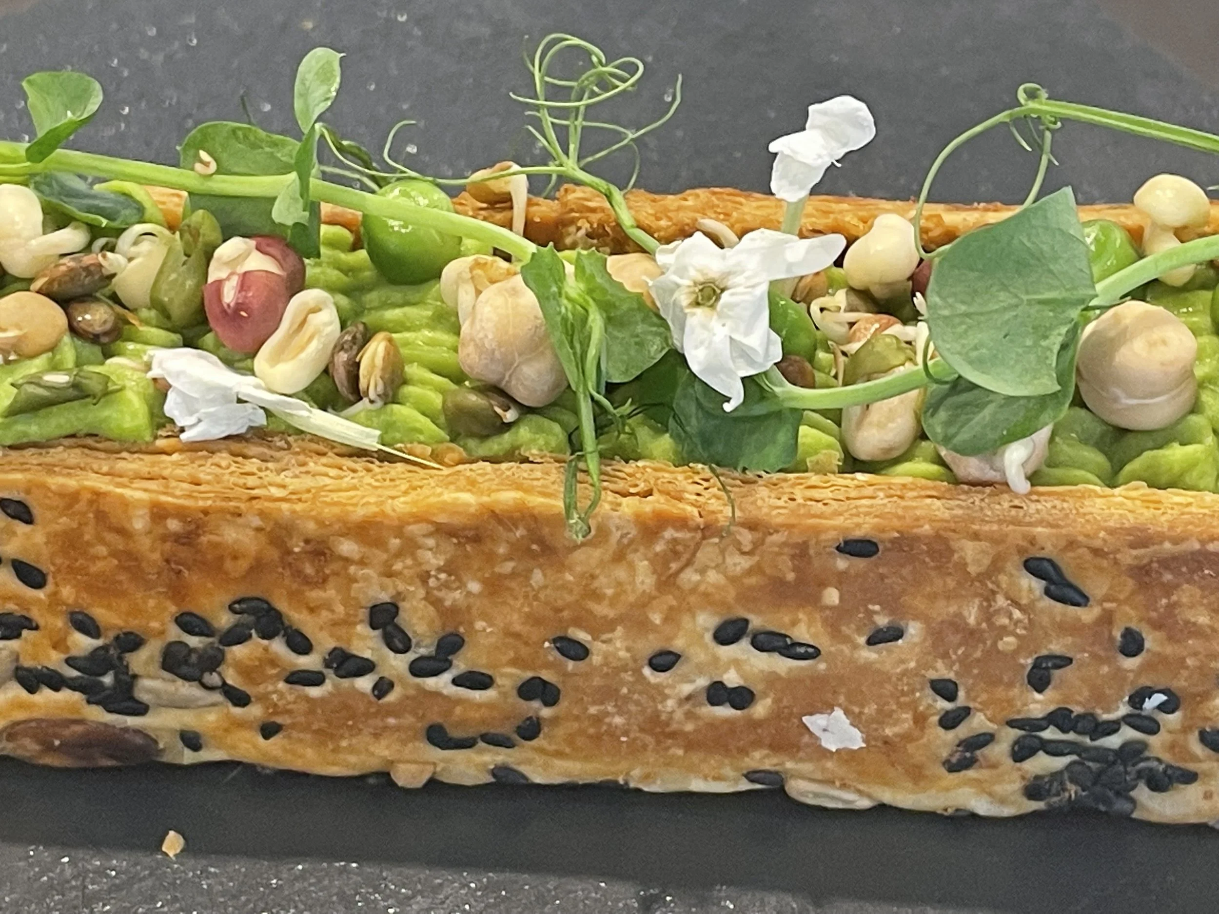 Terrine de poisson avec graines de sésame noir, avocat, pois chiches, micro-pousses et fleurs blanches garnie d'une garniture verte.