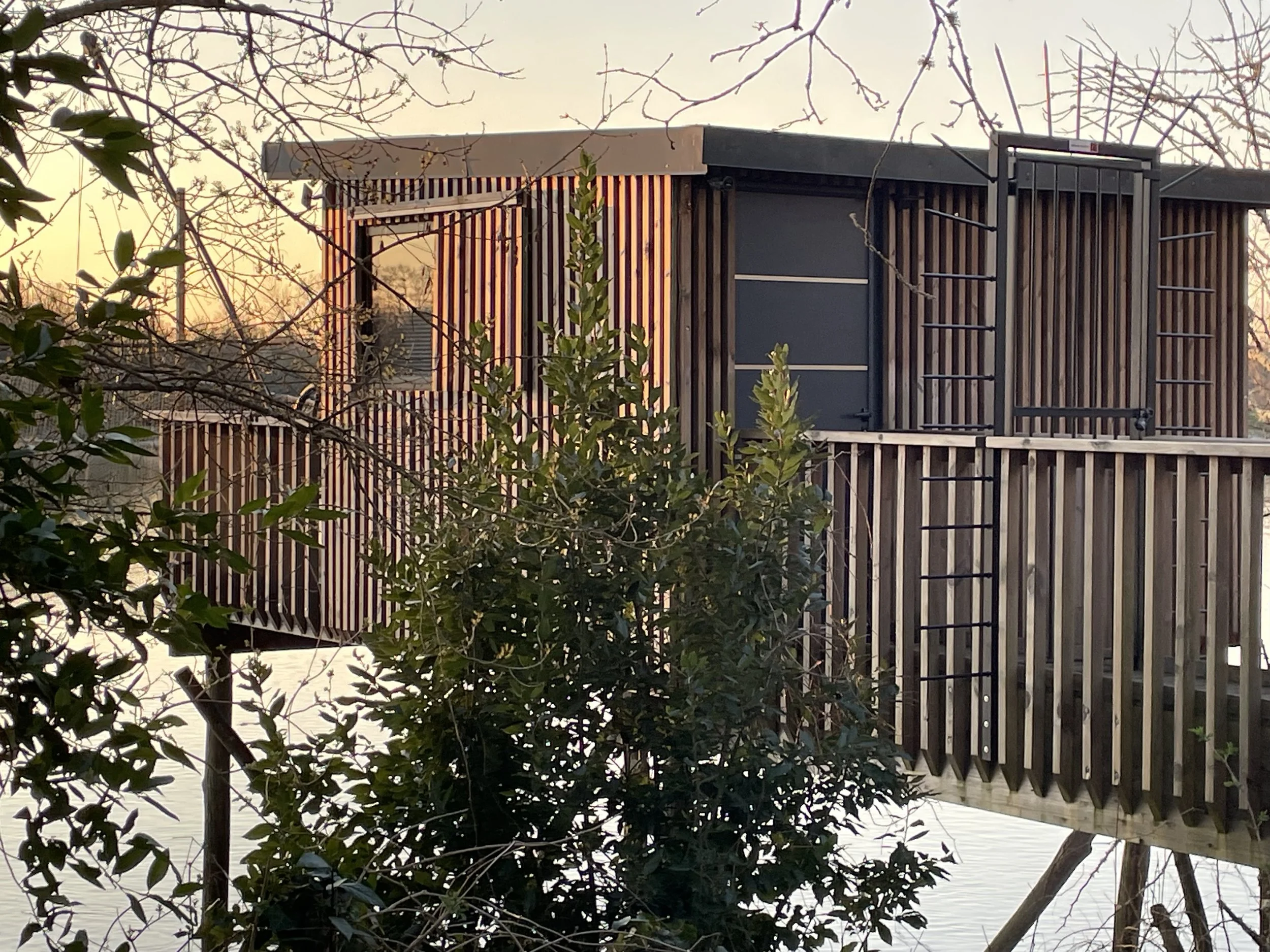 Une maison en bois construite sur pilotis au bord de l'eau, entourée d'arbres sans feuilles, avec un coucher de soleil en arrière-plan.