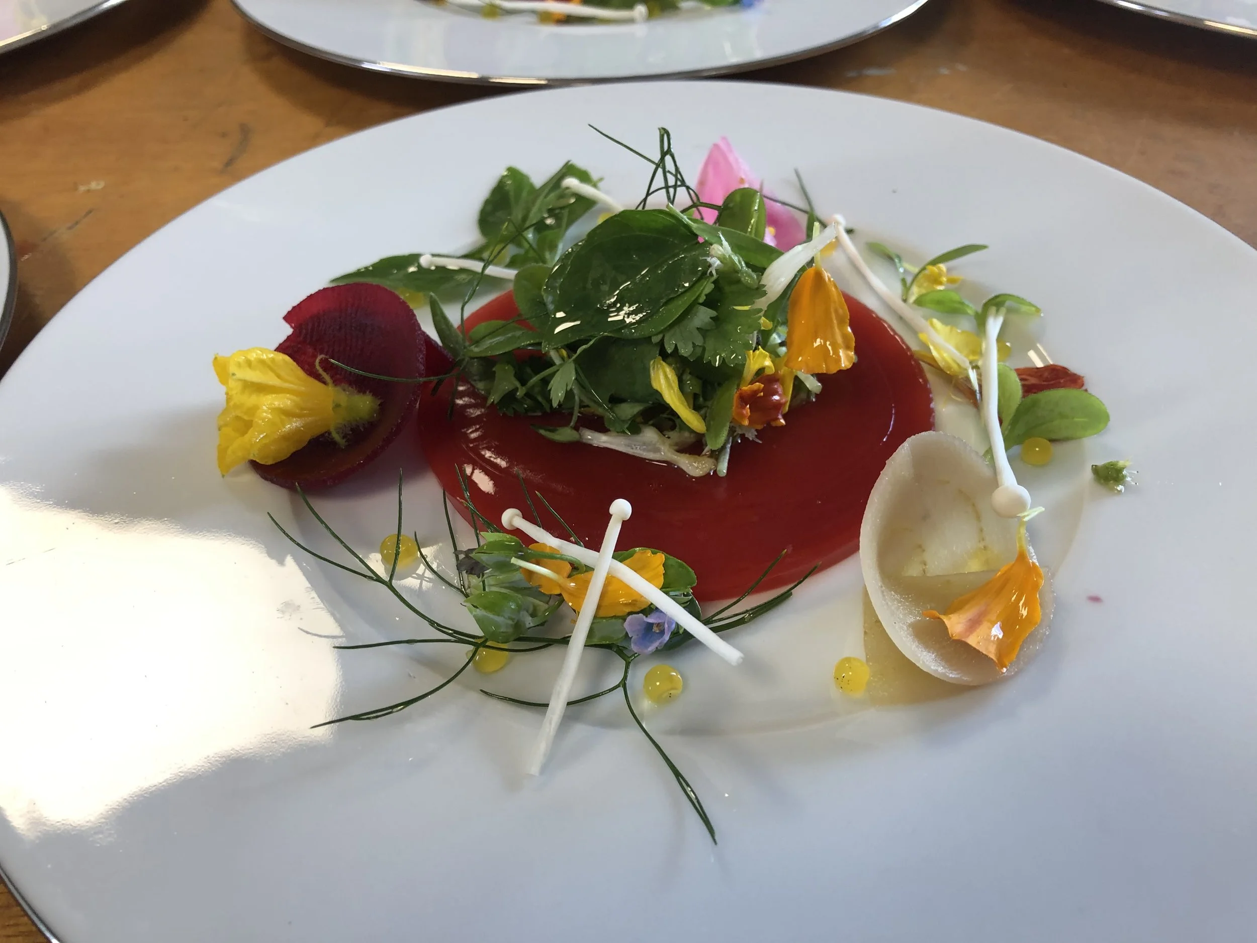 Assiette gastronomique avec des fleurs comestibles, des micro-pousses, une sauce rouge, et des éléments décoratifs colorés.