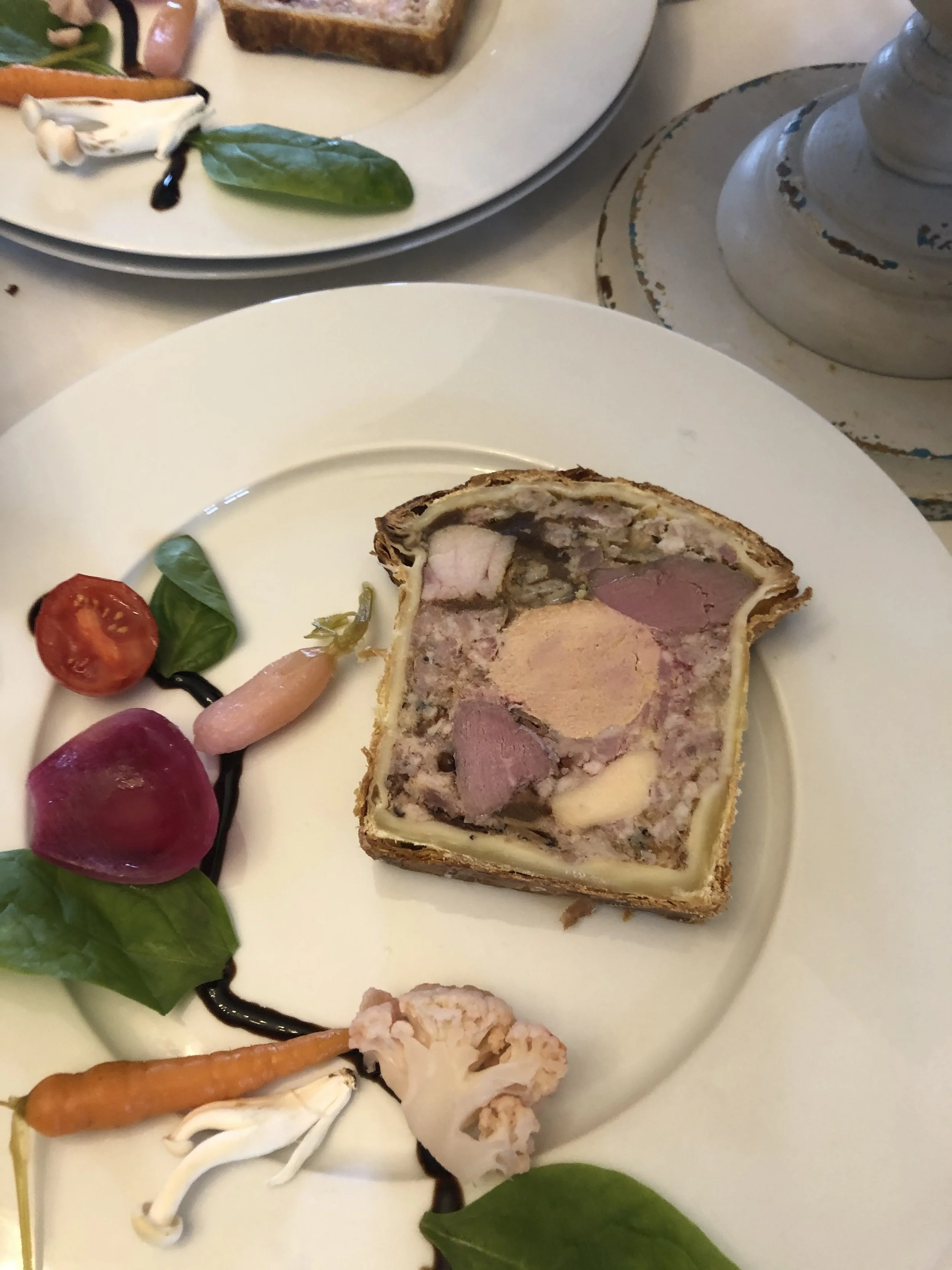 Assiette de pâté en croûte accompagnée de légumes entiers comme tomate, radis, carotte, chou-fleur, et des feuilles de basilic, décorée avec une sauce balsamique.