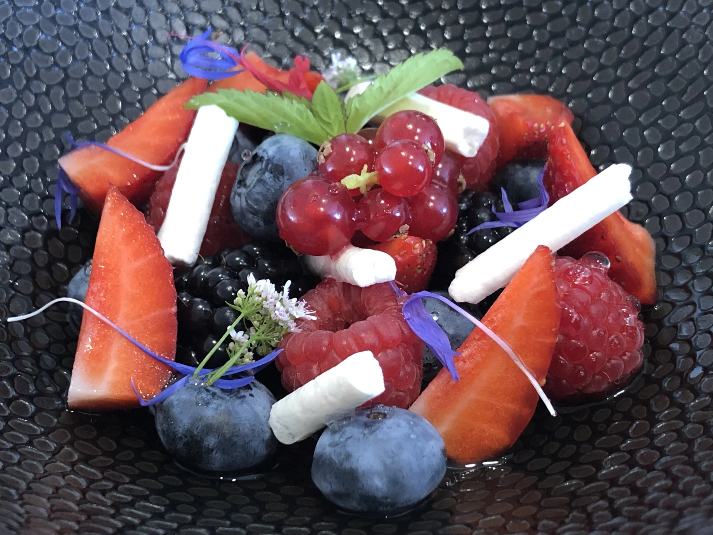 Salade de fruits avec fraises, myrtilles, groseilles, mûres, avec des fleurs violettes, feuilles de menthe et morceaux de fromage blanc.