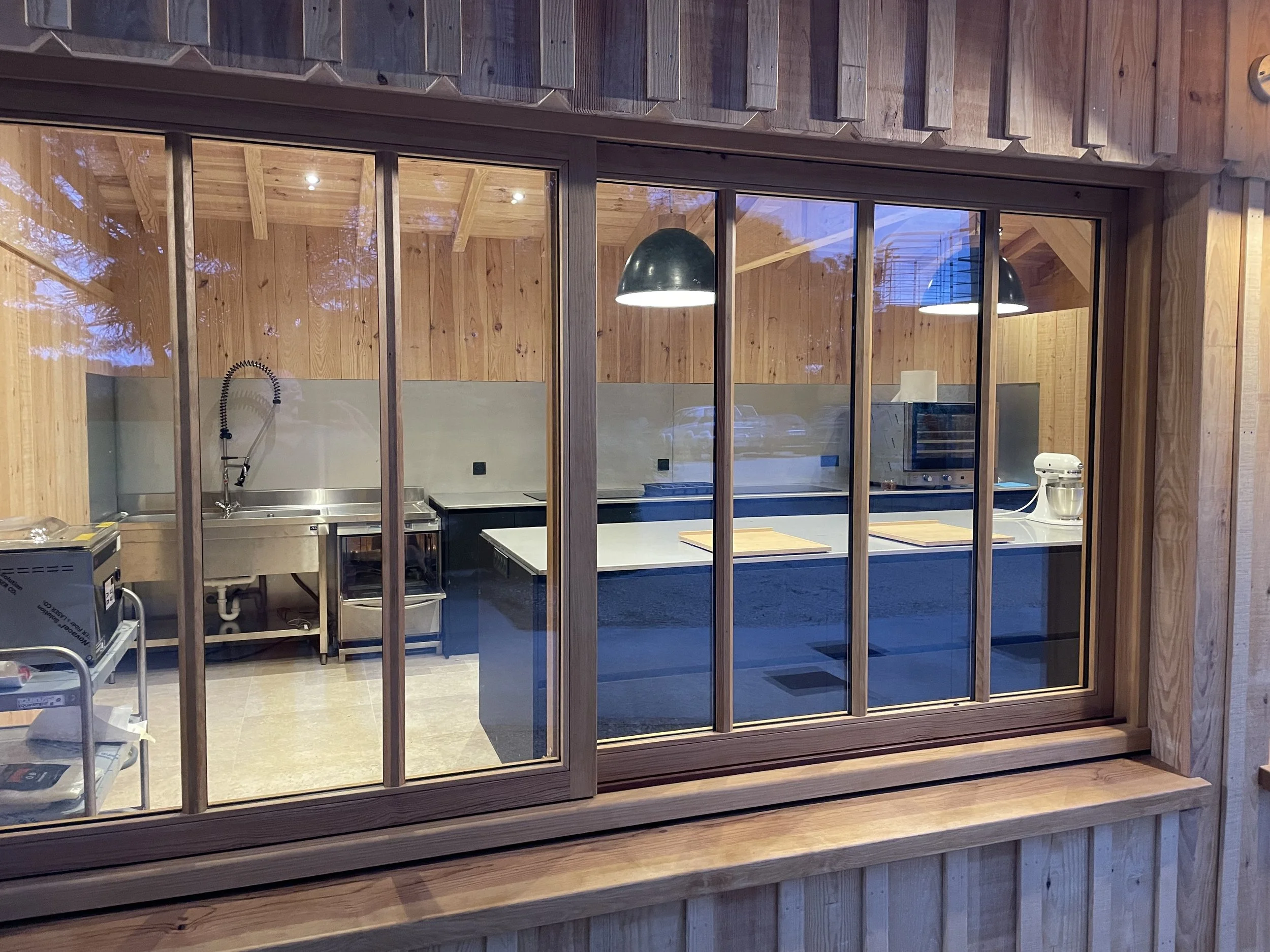 Cuisine moderne visible à travers une fenêtre en bois avec plusieurs lampes suspendues, évier, four et autres appareils électroménagers.