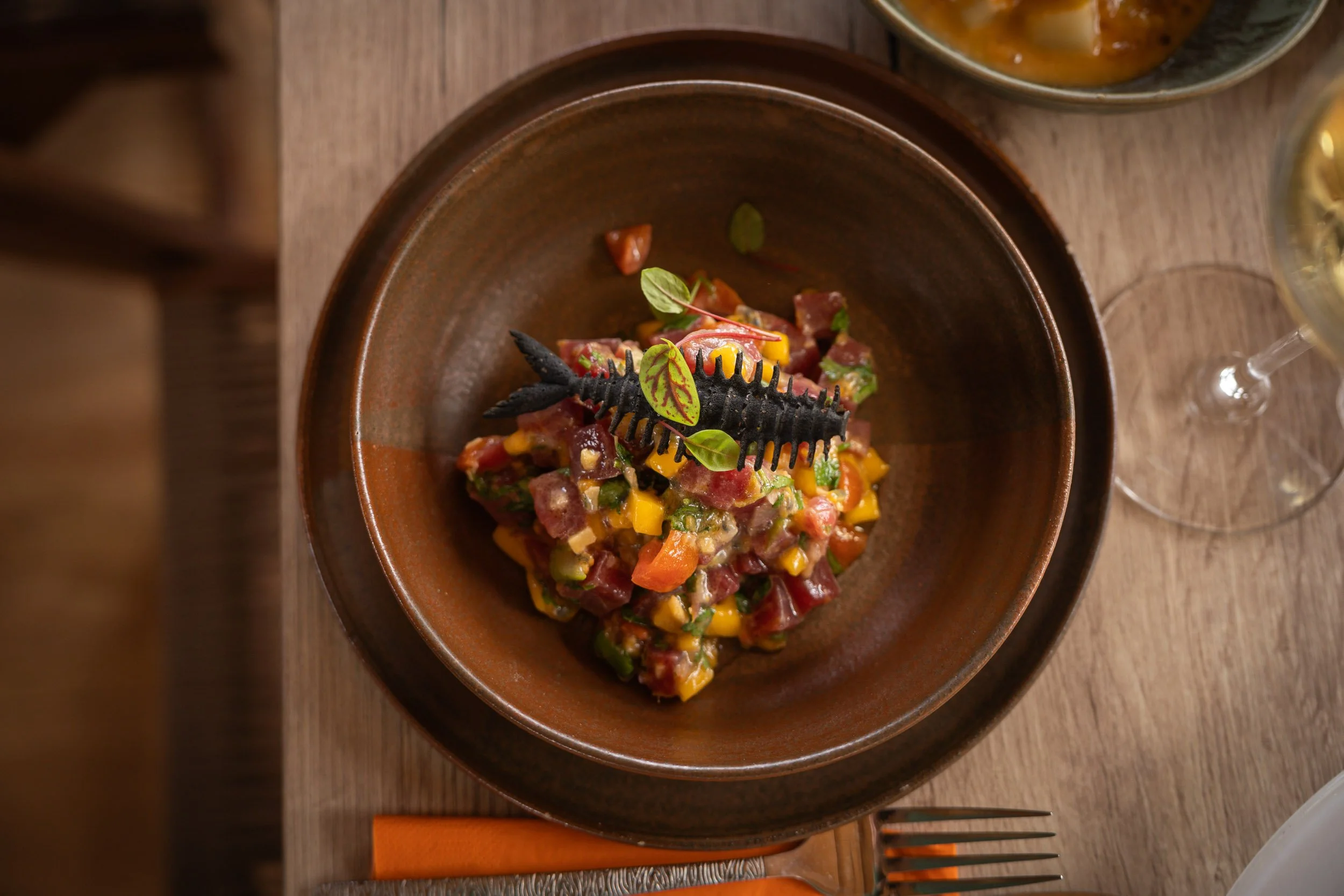 Salade de tartare de poisson avec un poisson en plastique noir sur le dessus, garnie de quelques feuilles vertes, servie dans un bol en terre cuite, avec un verre de vin blanc à côté.