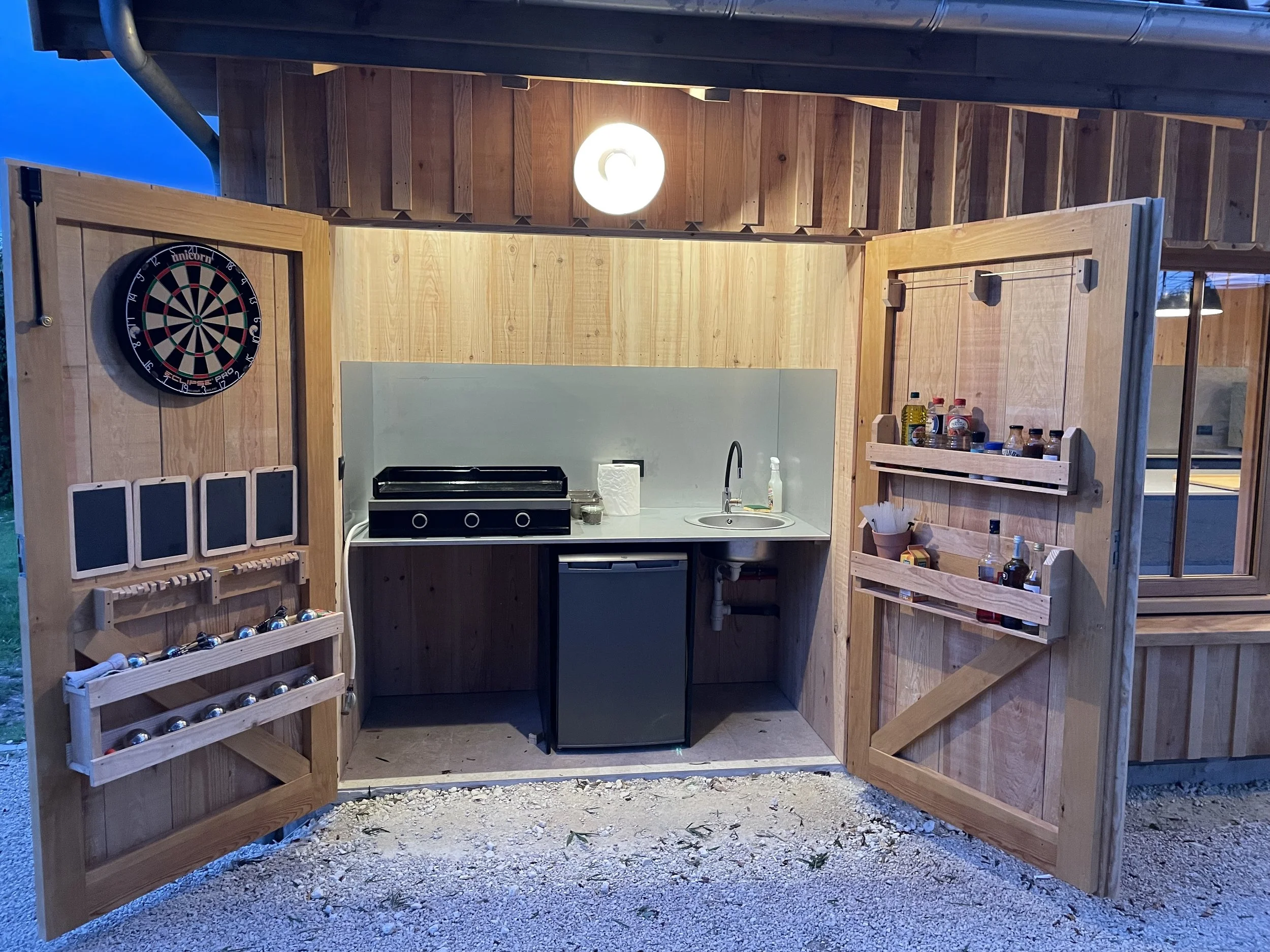 Un mini-bar en bois avec une porte ouverte, comprenant un tableau de fléchettes à gauche, un petit four électrique, un évier, une petite réfrigération et des étagères avec des épices et des bouteilles