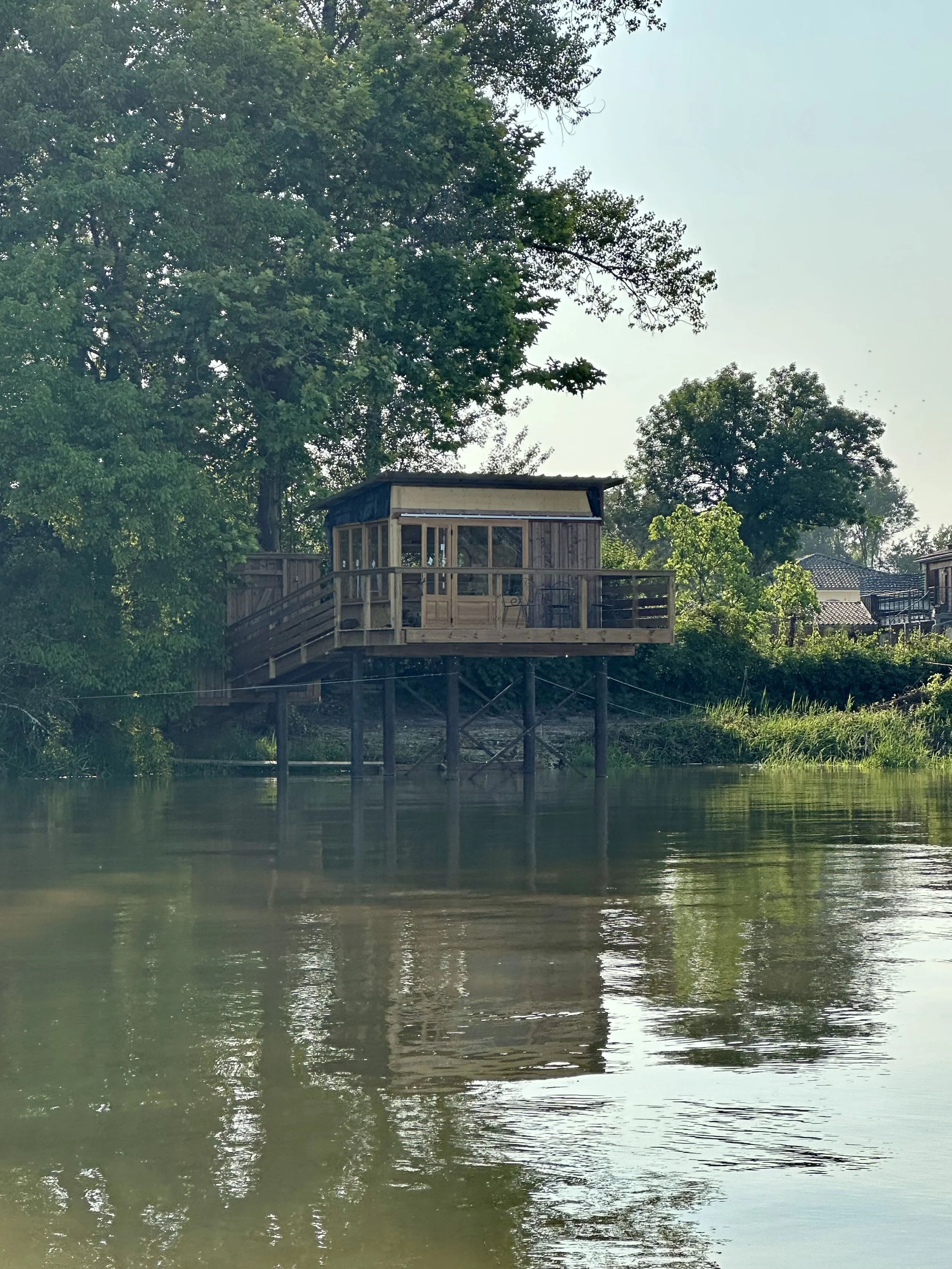Une cabane en bois construite sur pilotis située au bord d'une rivière, entourée d'arbres verts et sous un ciel clair.