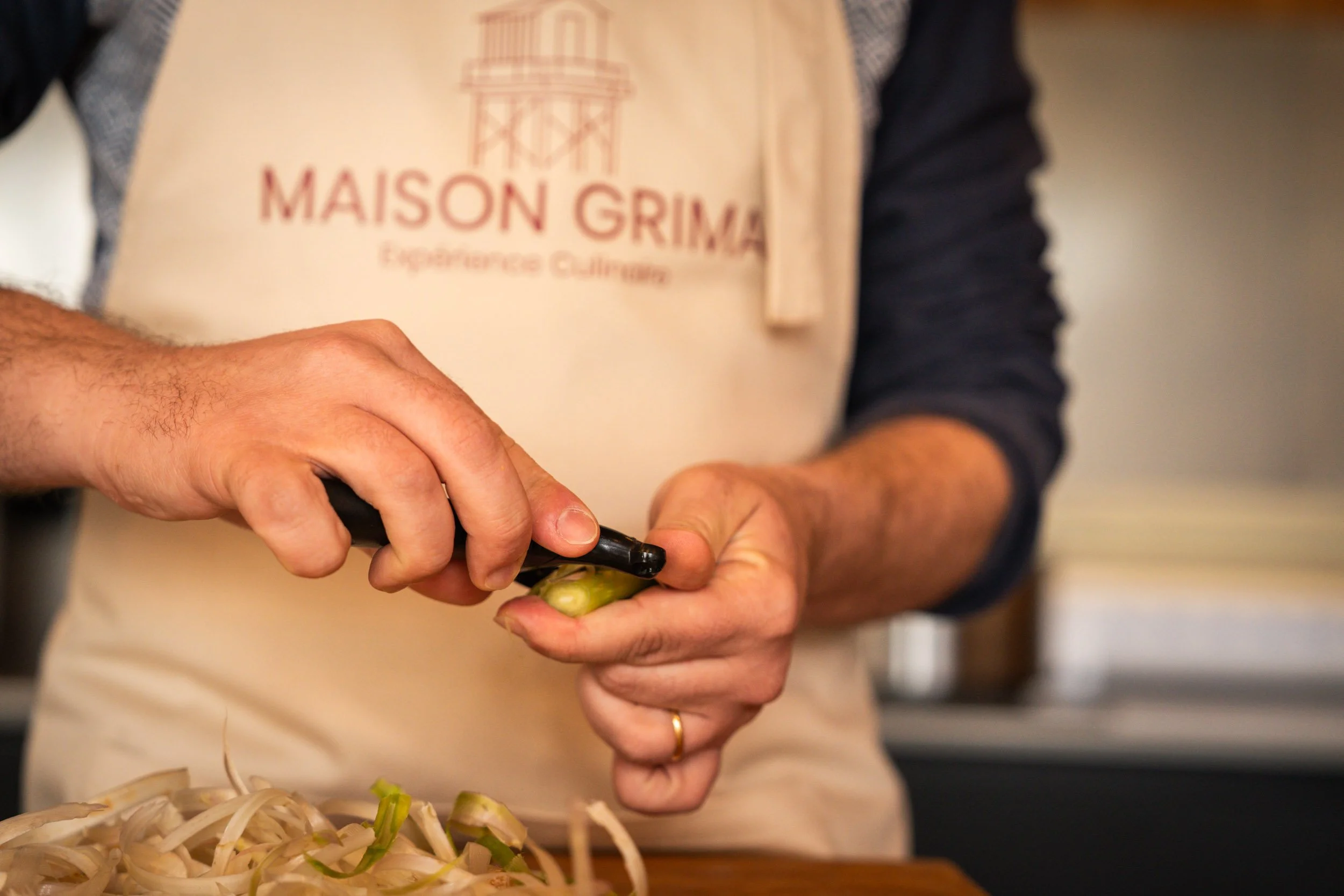 Une personne qui coupe un légume avec un couteau de cuisine, portant un tablier beige avec l'inscription 'Maison Grima'.