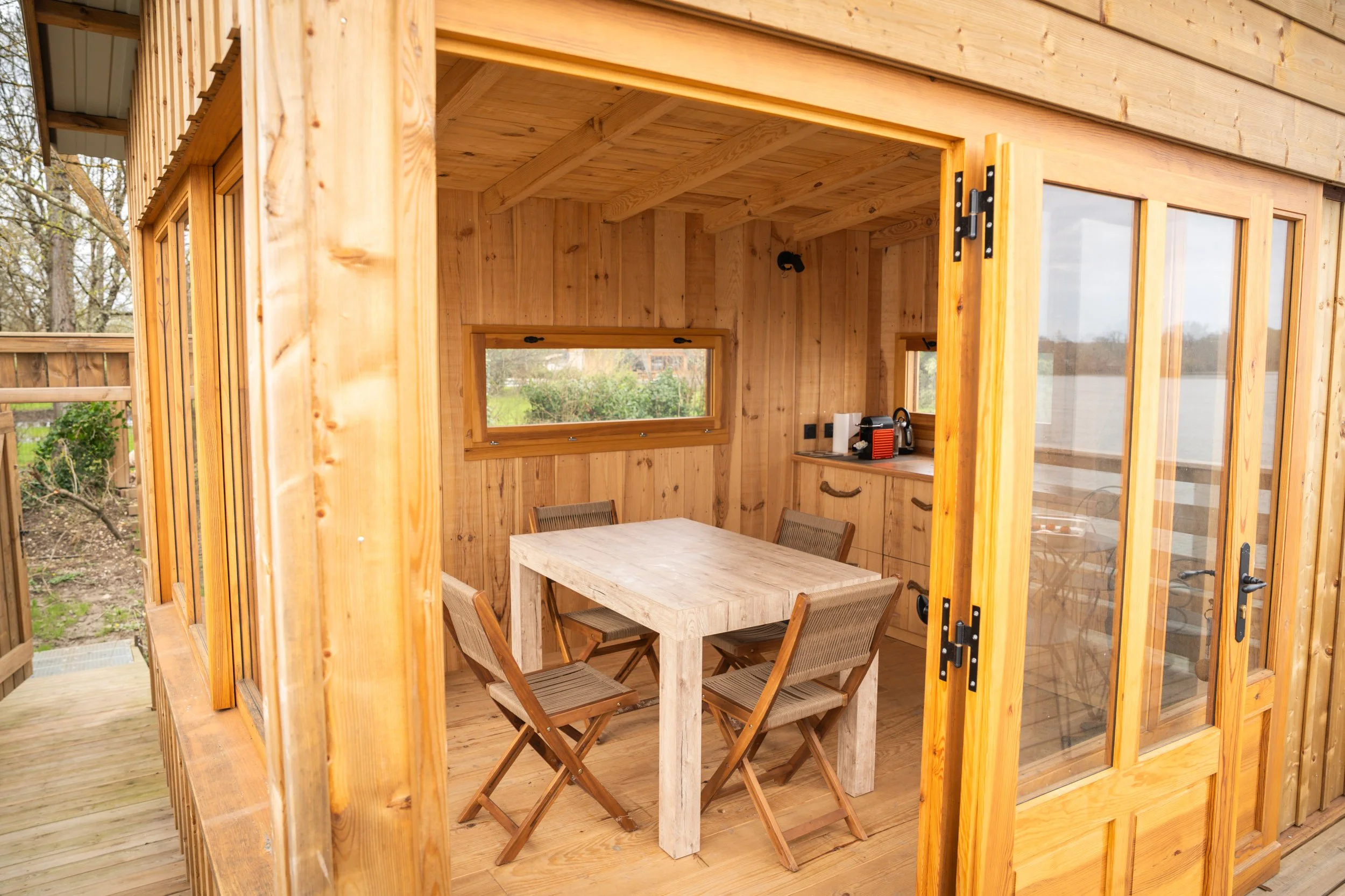 Une petite cabane en bois avec une table en bois et quatre chaises en bois à l'intérieur, équipée d'une petite kitchenette avec un grille-pain, une bouilloire et quelques papiers.