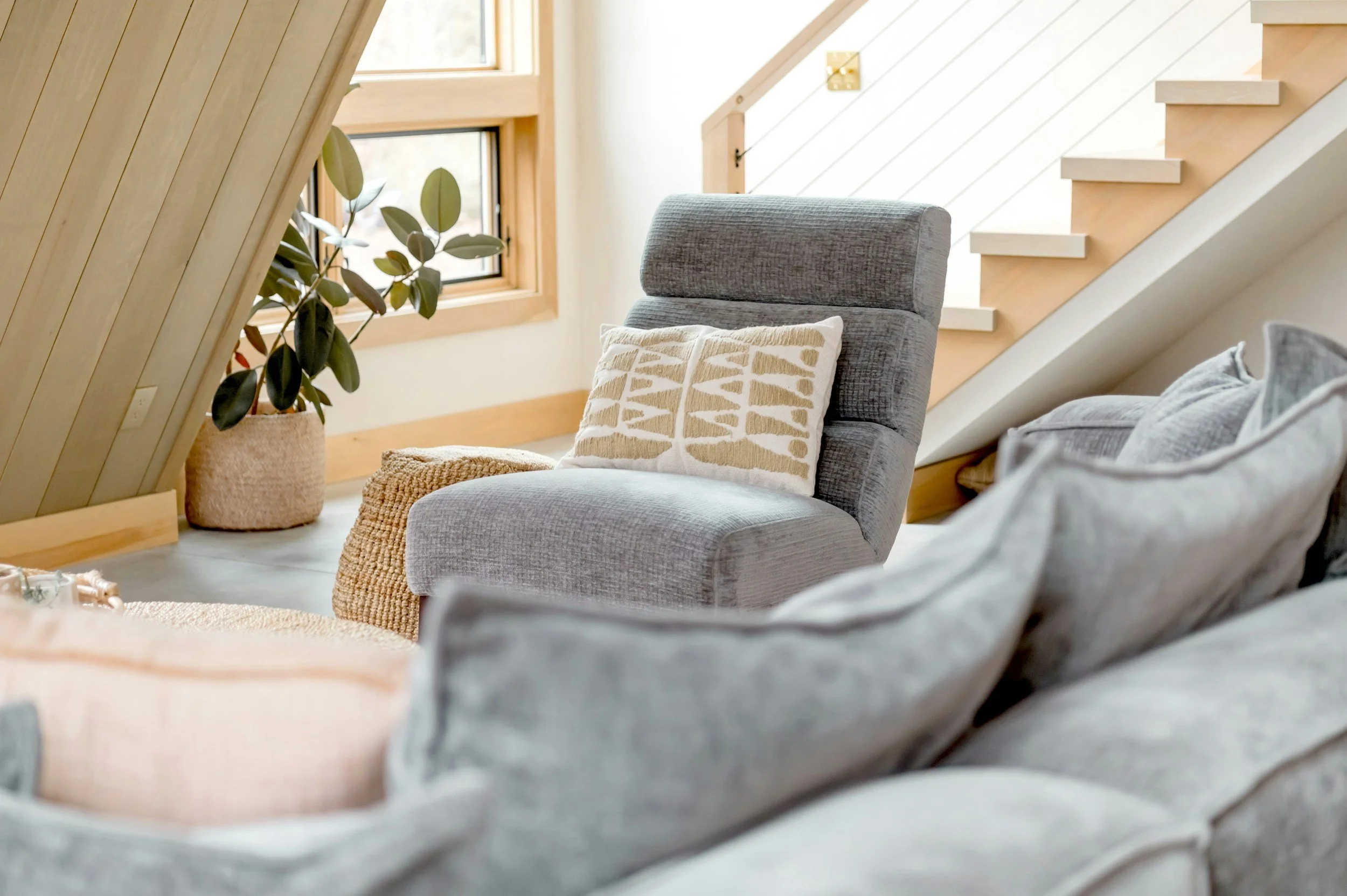 A cozy living room corner with a light gray armchair featuring a decorative pillow, a plant in a woven basket, and a staircase with wooden steps and a white railing in the background.