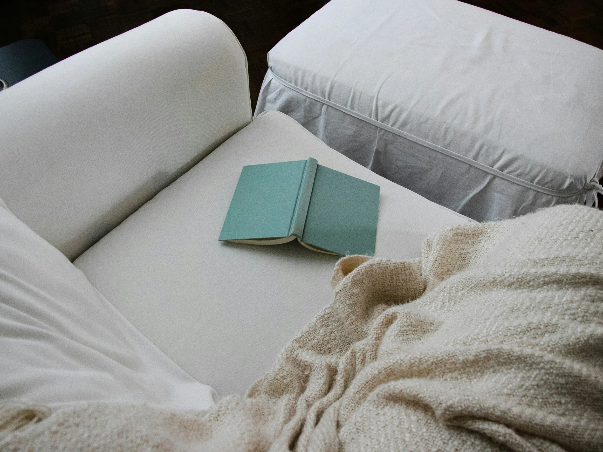 A person sitting on a white sofa with an open blue book on their lap, a knitted blanket covering their legs, and a white cushion and mattress in the background.