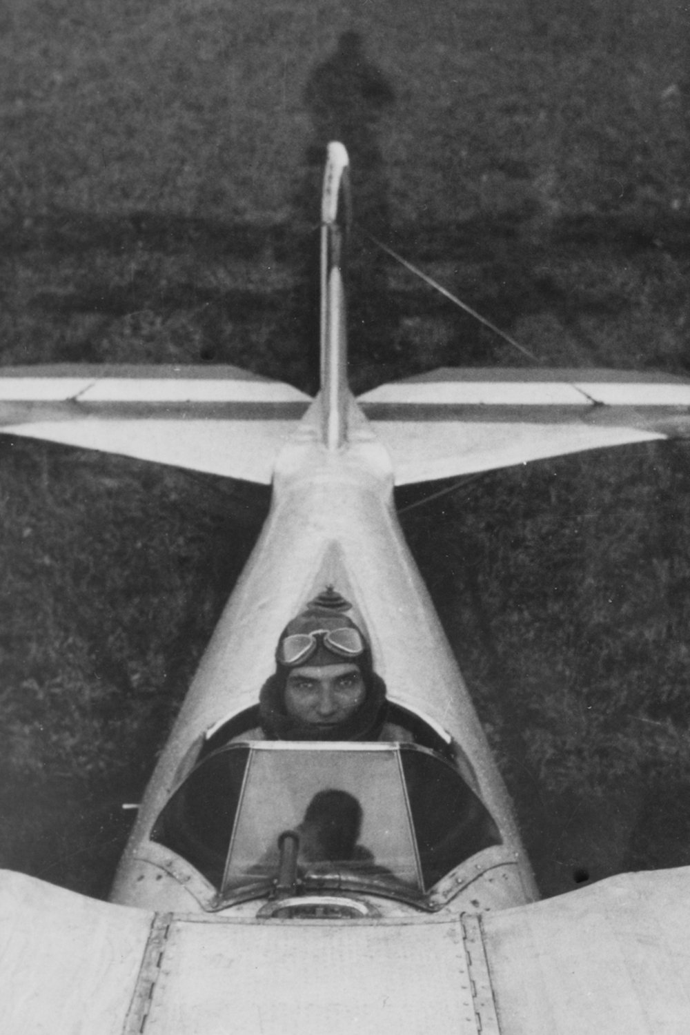 Black and white aerial view of a woman pilot in the cockpit of a fighter jet, wearing goggles and a scarf, with the jet's wings extending on either side.