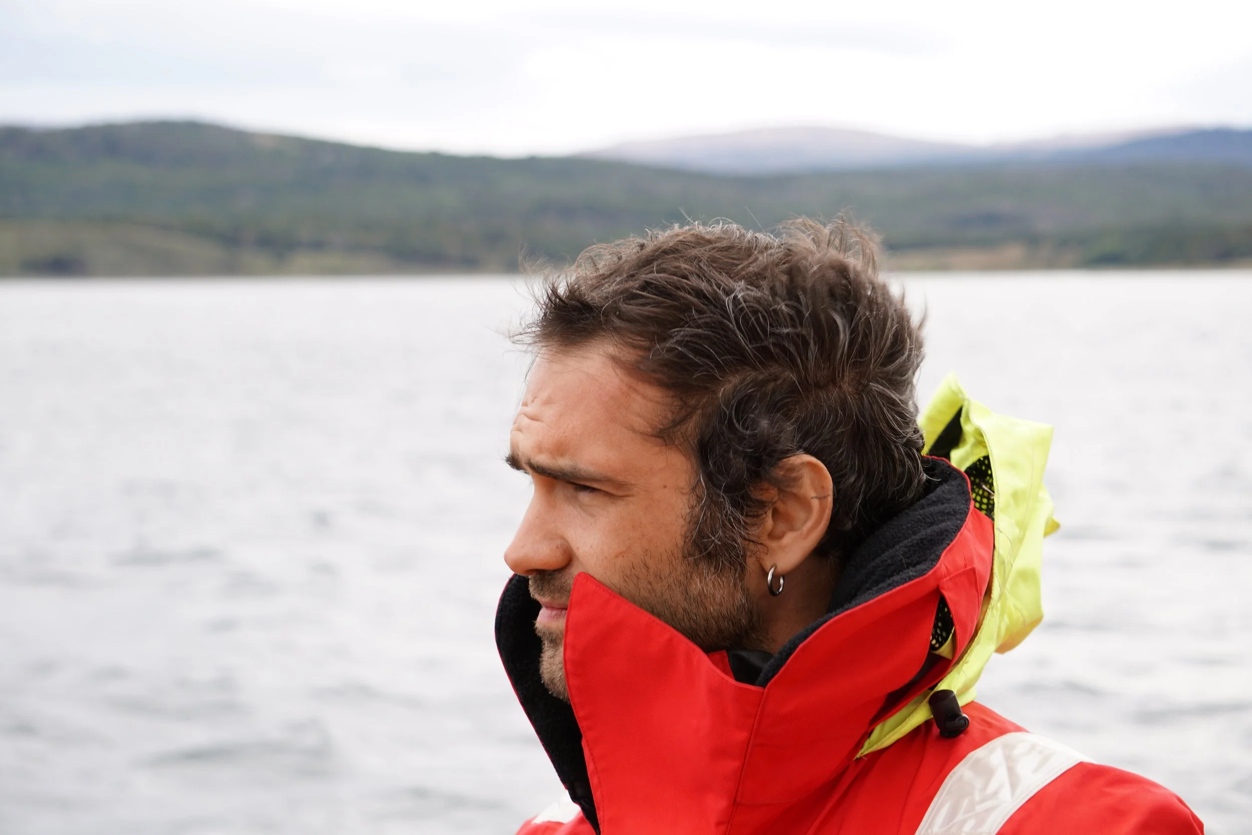 Manfredi Calamai, a man with dark hair and a beard wearing a red and yellow waterproof jacket, standing by a body of water with mountains in the background.