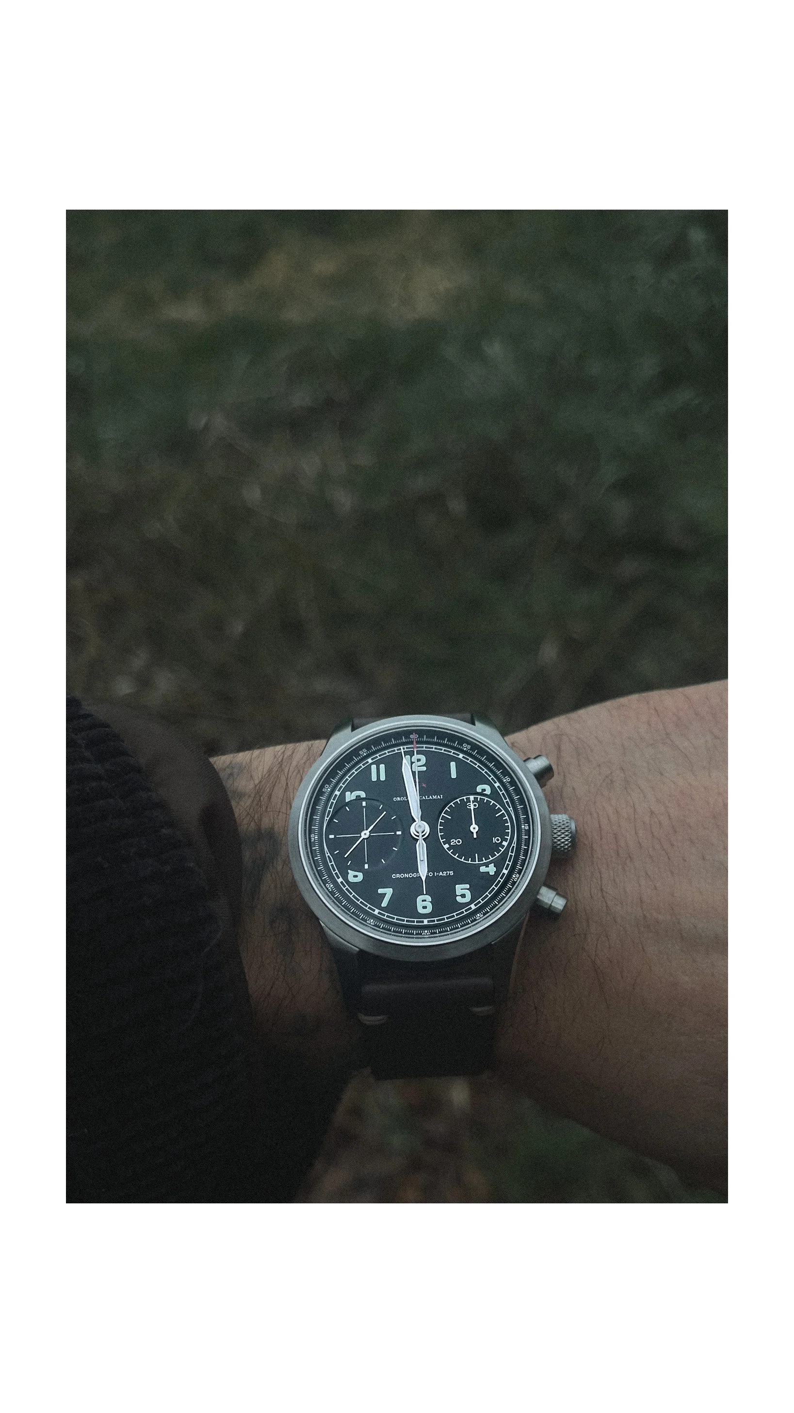 A person's wrist wearing a black chronograph watch with a black dial and white markings, with an outdoor grassy background.