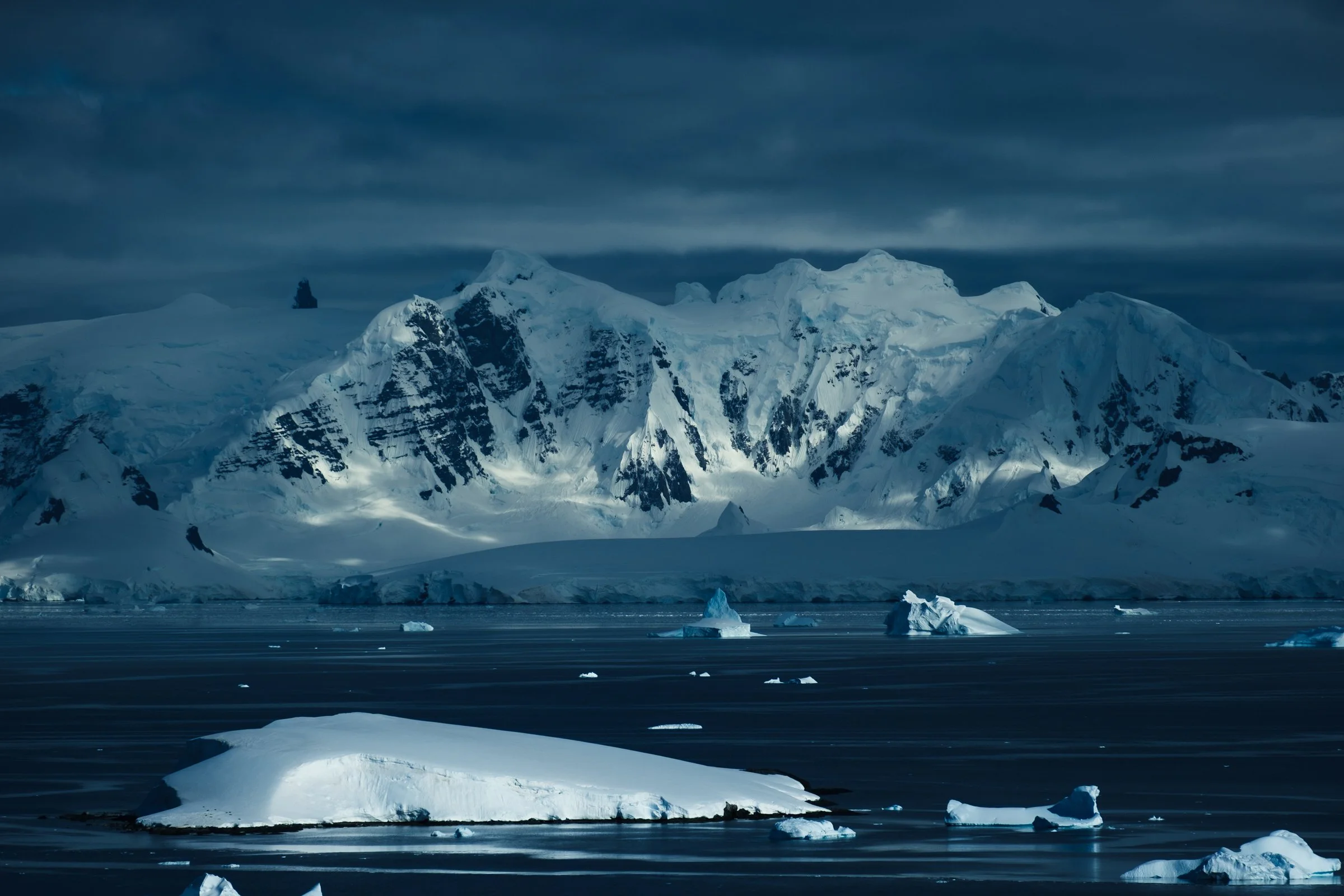Snow-covered mountains and icebergs floating in dark ocean under cloudy sky in Antarctica.